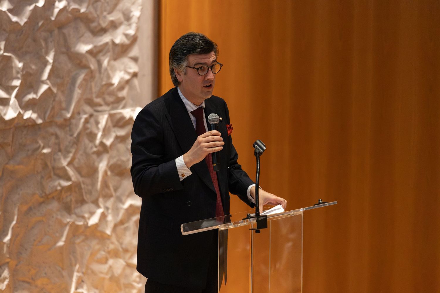 A man in a dark suit speaks at a clear podium with a microphone in a modern venue with textured walls.