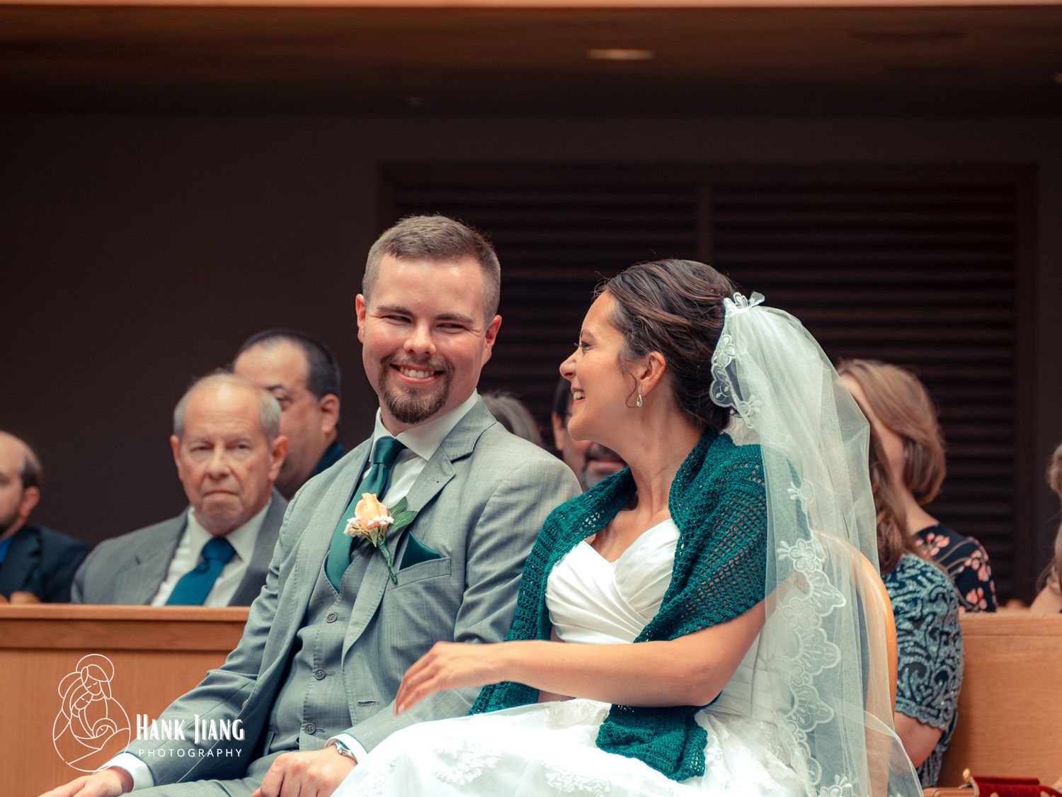 Smiling bride in white veil and groom in gray suit sharing a joyful moment during wedding ceremony.