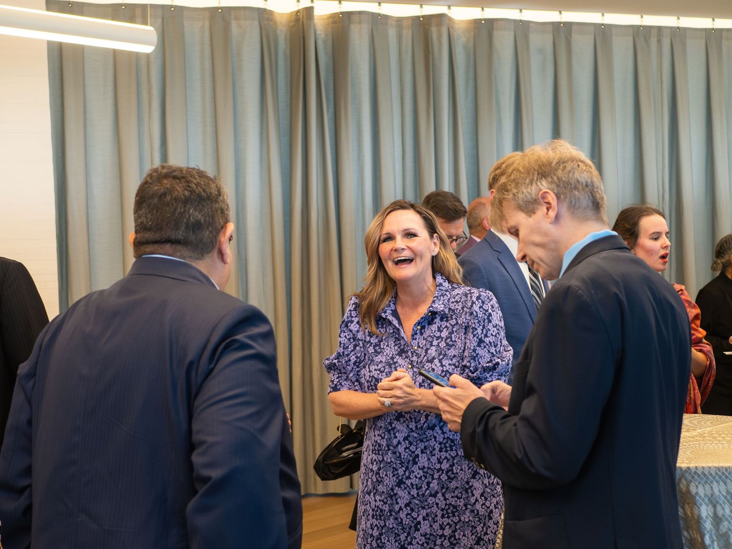 Professionals networking and laughing at a corporate event in a modern venue with teal curtain backdrop.