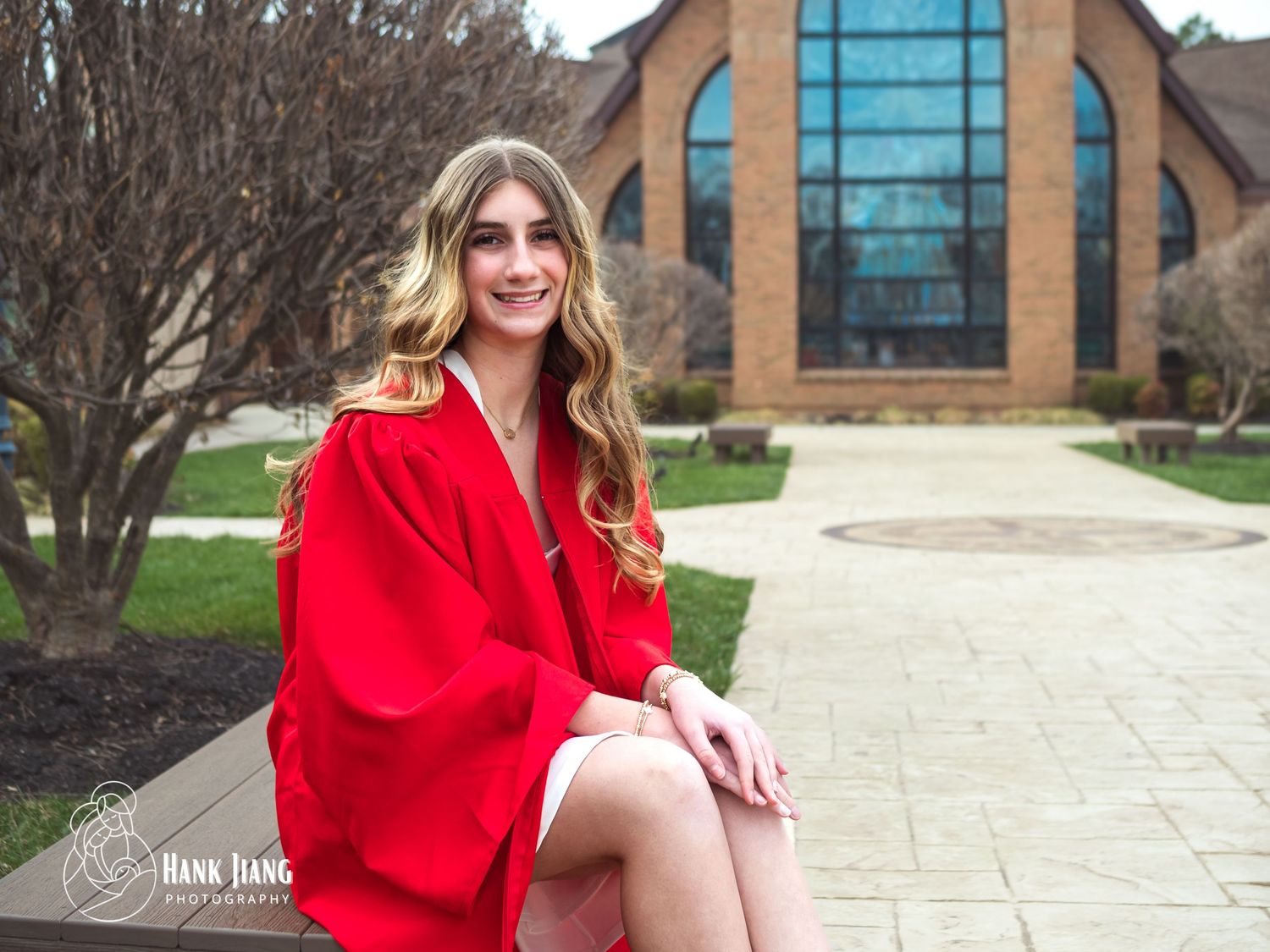 Young woman in red graduation robe sitting outdoors near a brick building, smiling for a senior portrait photo.