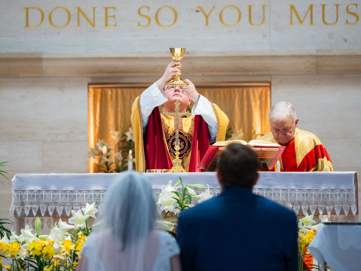 Catholic priest elevating the chalice during wedding Mass with bride and groom at the altar.