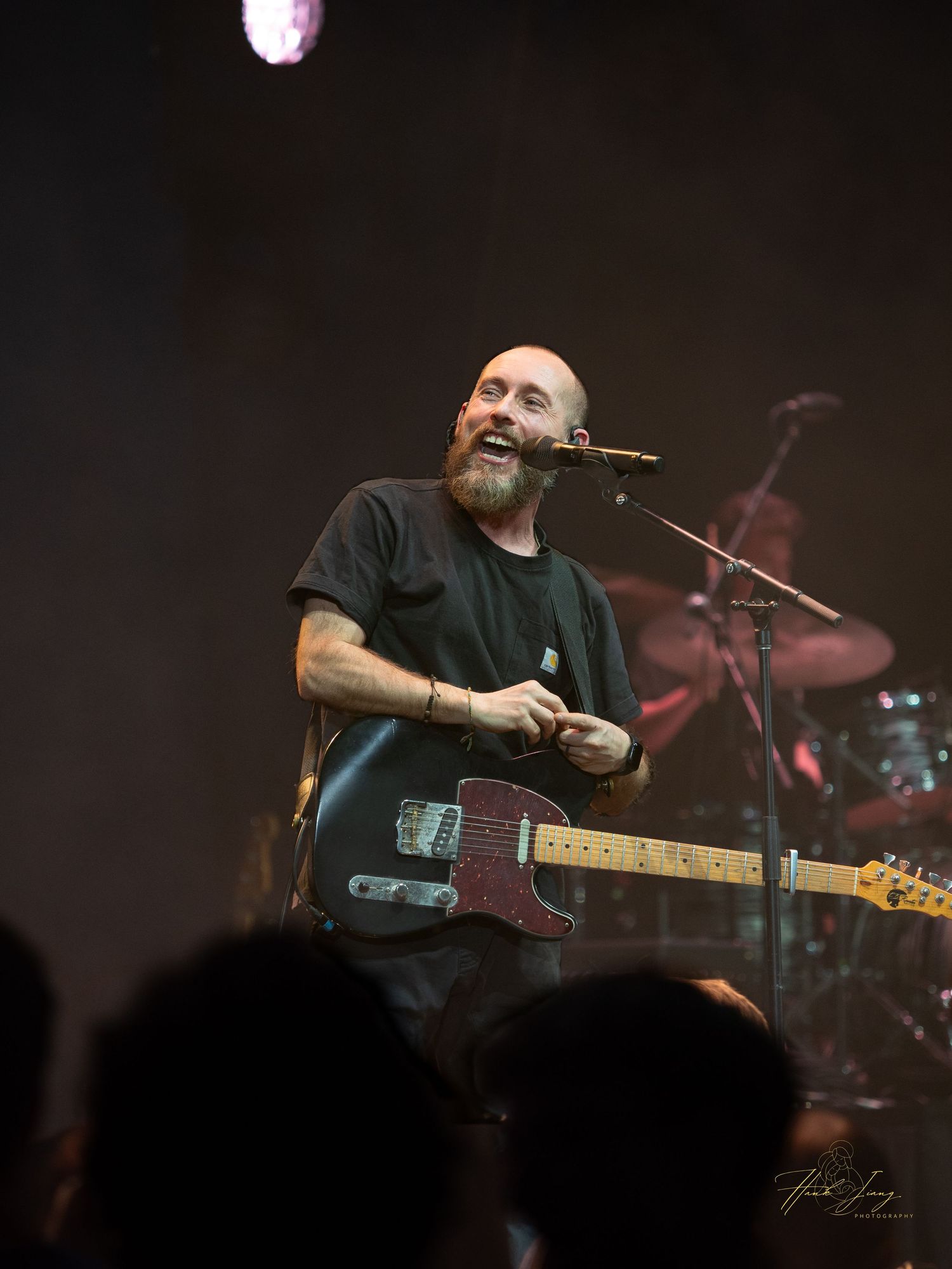 Smiling bald bearded musician playing a sunburst Telecaster guitar on a smoky concert stage before a live audience.
