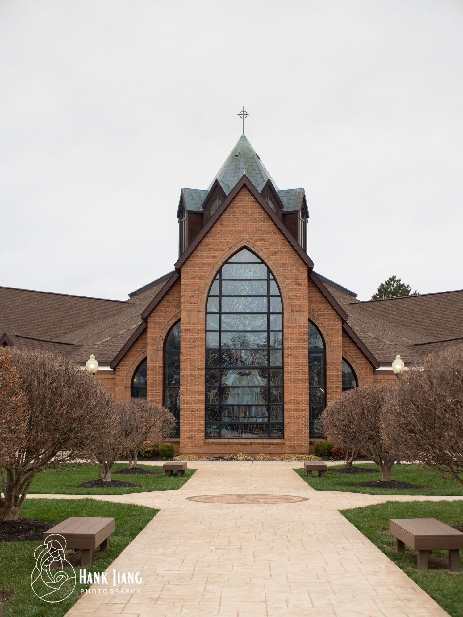 Brick church with gothic arched windows, cross steeple, and stone pathway surrounded by bare trees on overcast day.