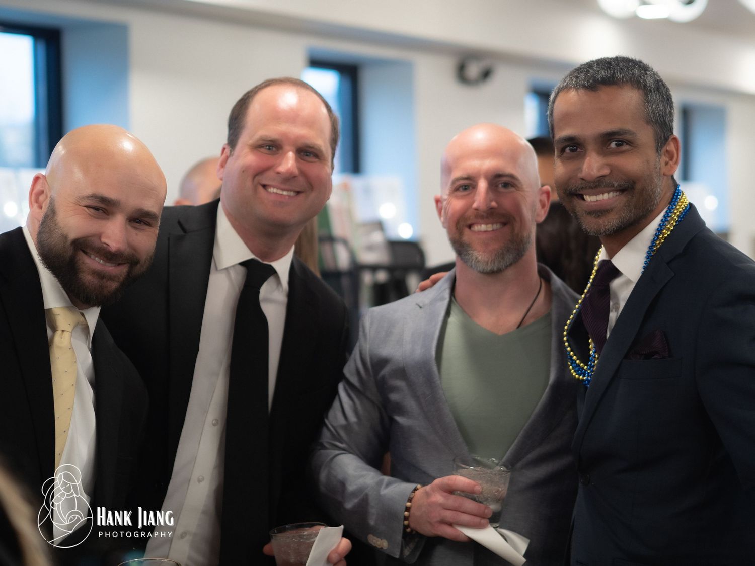 Four men in formal attire smiling together at an indoor social event or party.