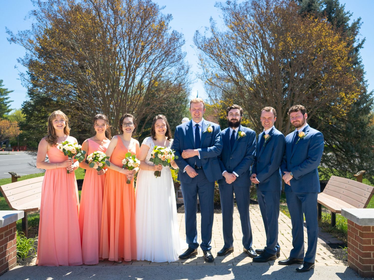 A wedding party in coral bridesmaid dresses and navy suits poses outdoors under sunny spring trees.