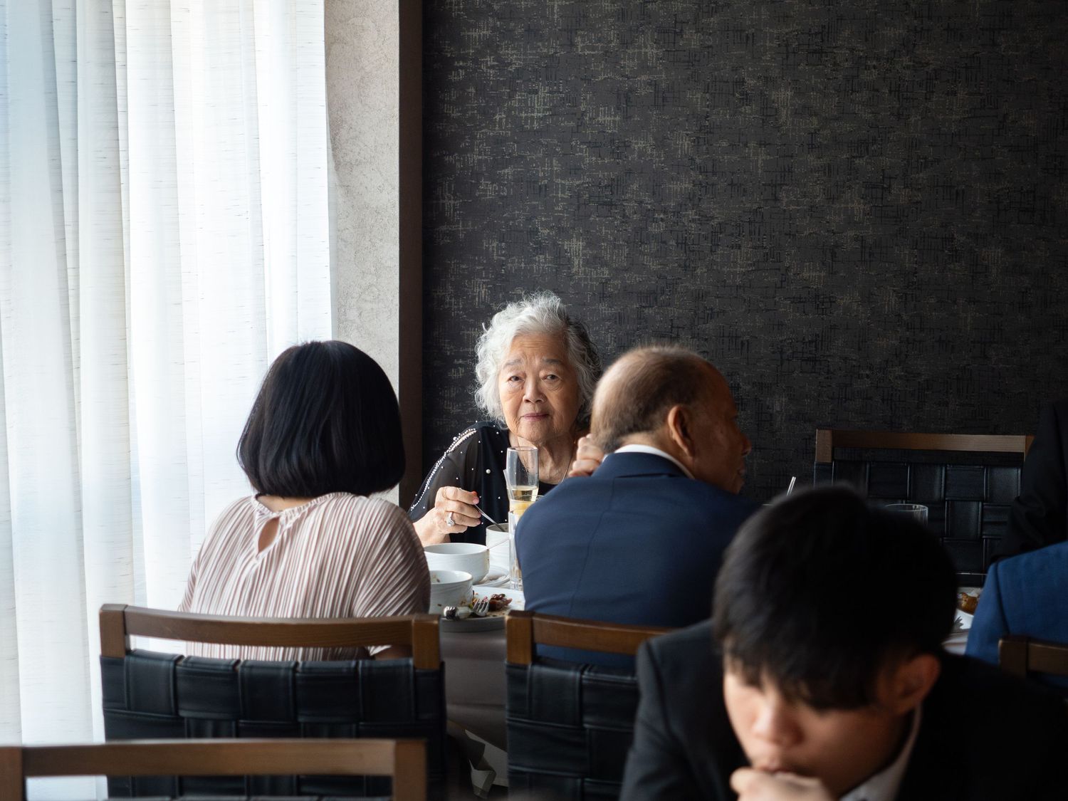 Elderly Asian woman smiling at a restaurant table with family members during a meal near a window.