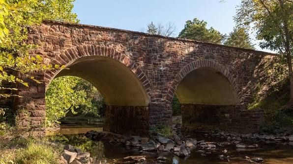 Historic red brick double-arch stone bridge spanning a shallow rocky creek surrounded by lush green trees.