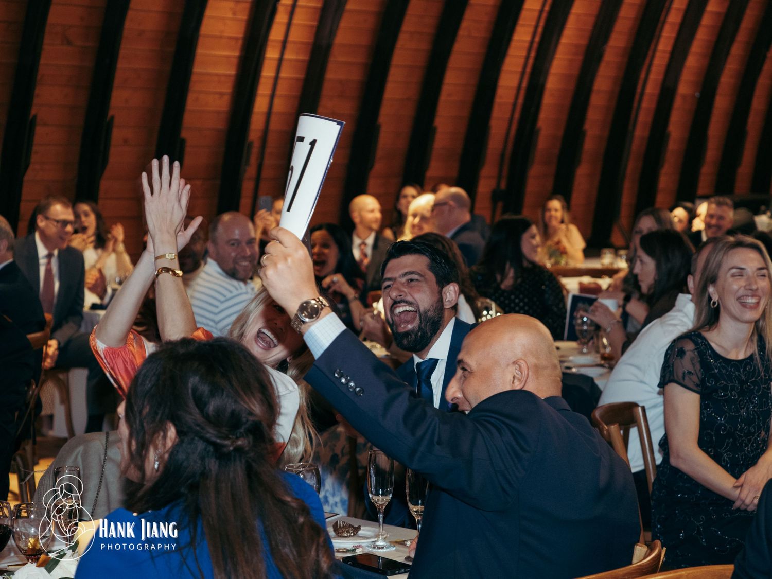 Guests at a formal gala event raise bidding paddles during a live auction inside an elegant wooden domed venue.