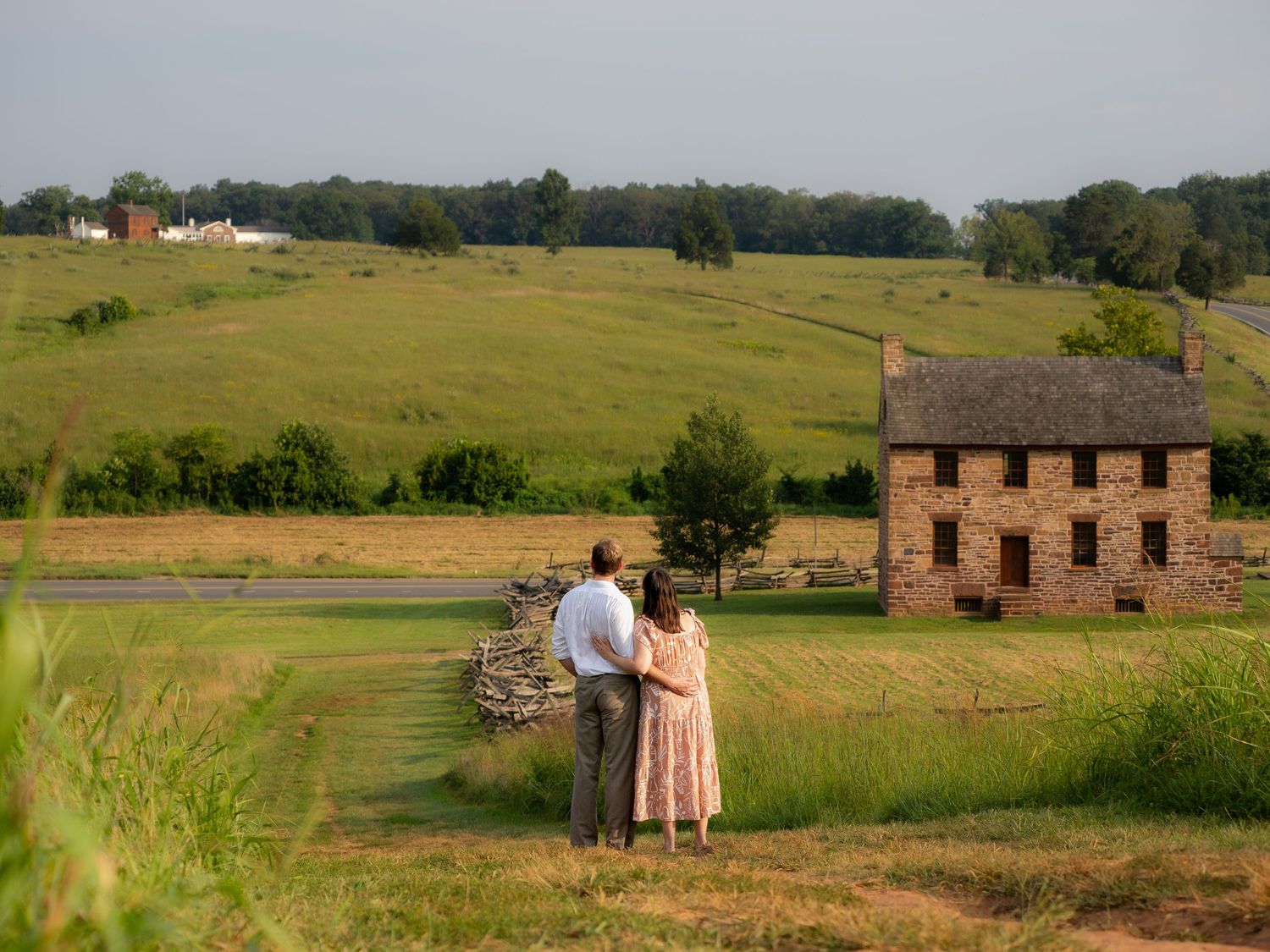 Couple stands embracing overlooking scenic green countryside with historic stone farmhouse.