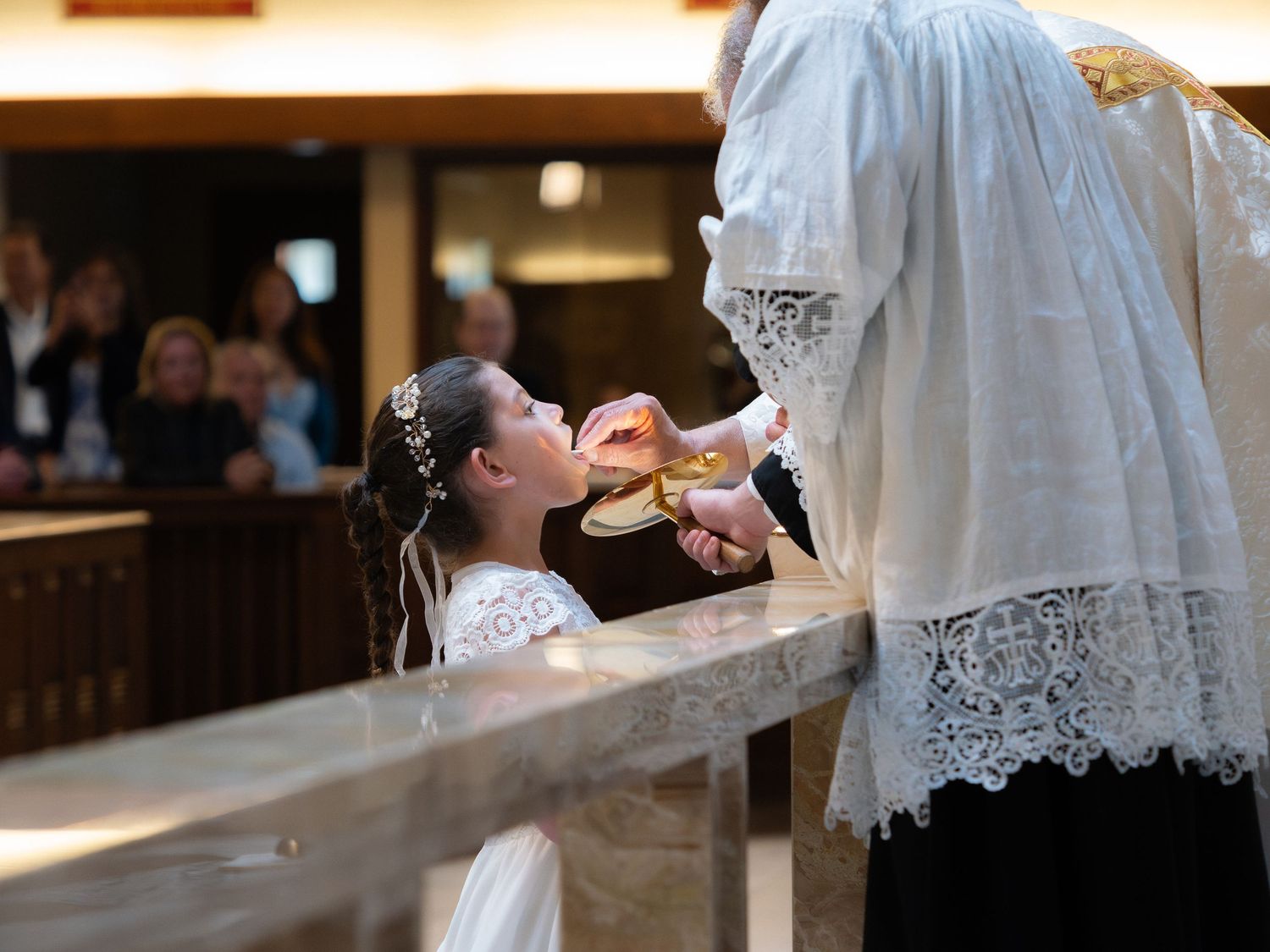 Young girl in white First Communion dress kneeling to receive the Eucharist from a priest at the altar rail.