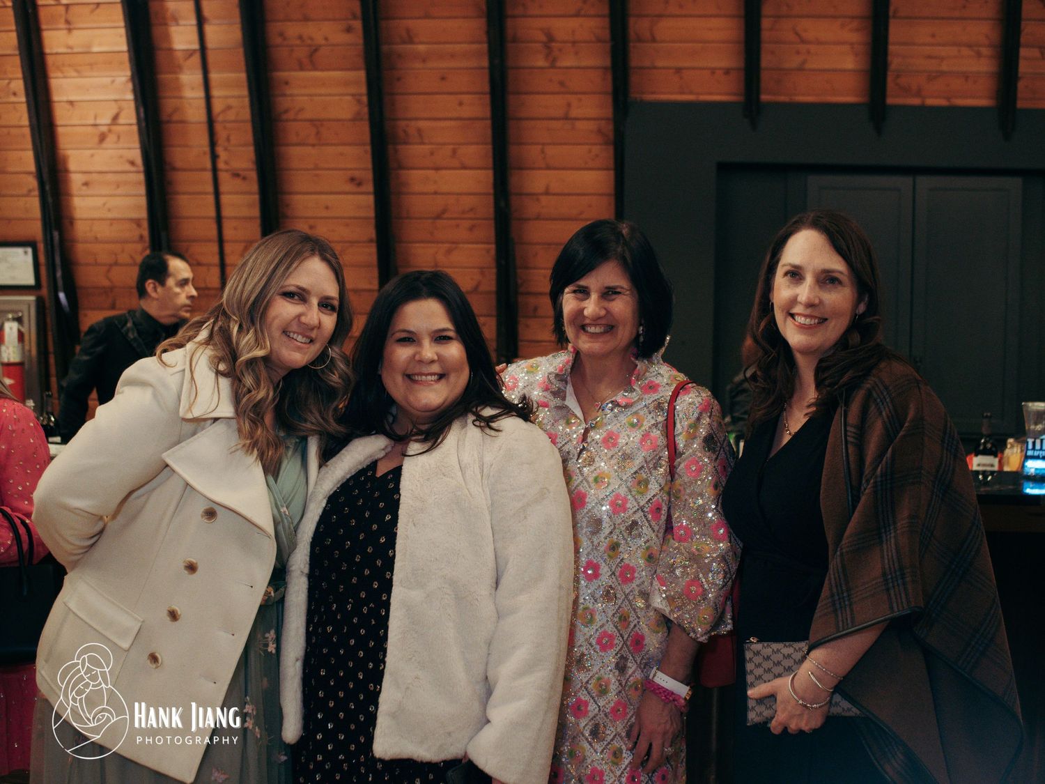 Four women smiling together at an indoor event, posing for a group photo in a rustic wood-paneled venue.