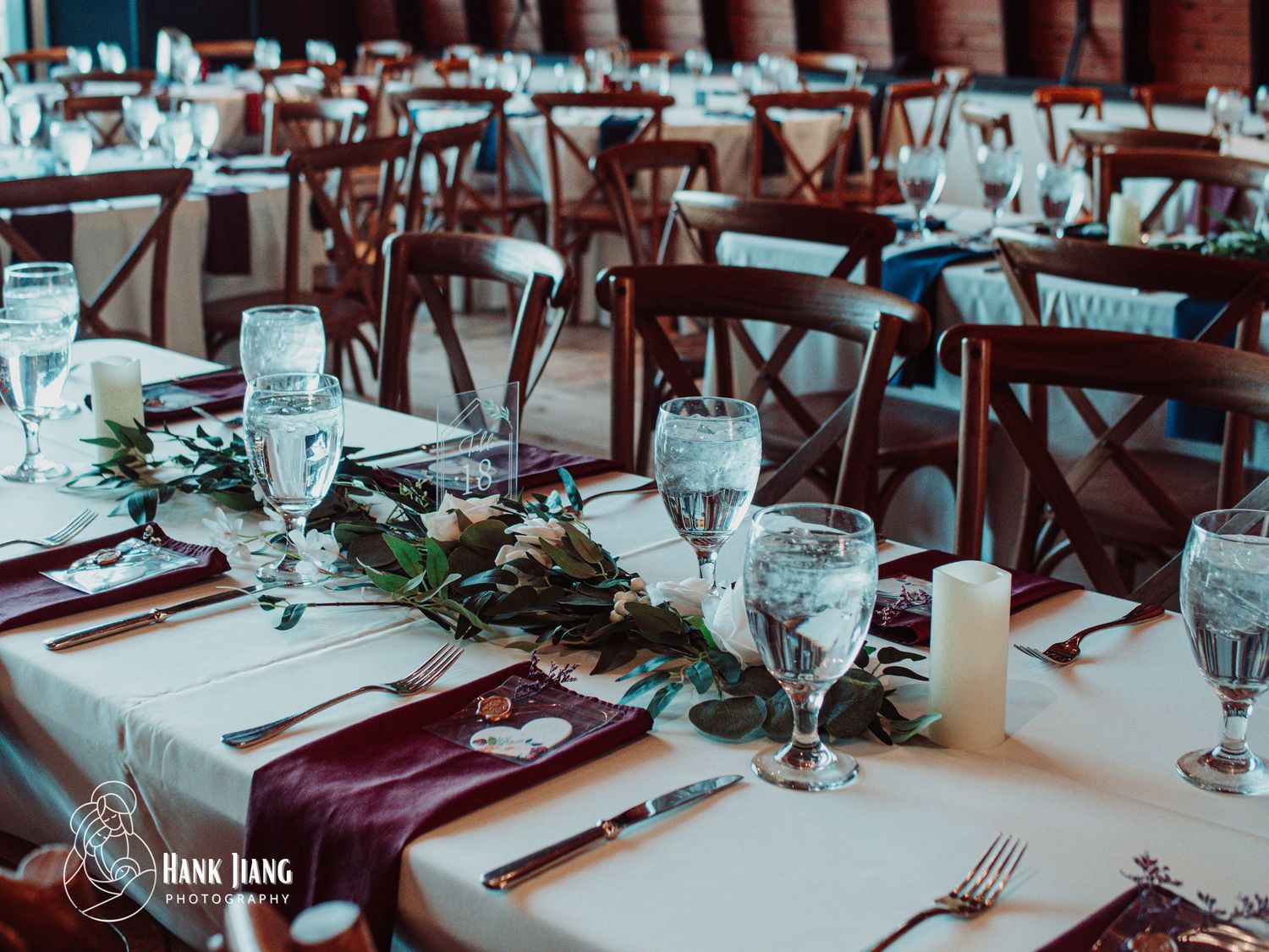Elegant wedding reception tables with burgundy napkins, greenery runners, candles, and crystal glassware in a rustic venue.