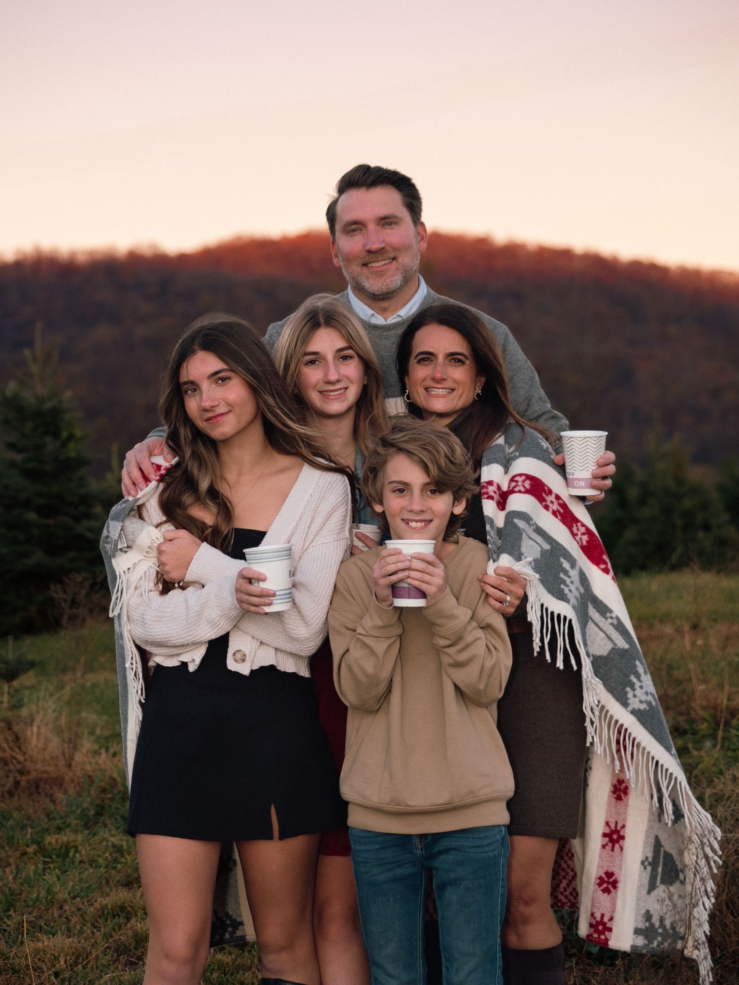Smiling family of five holding mugs at golden hour with a scenic mountain and evergreen tree backdrop.