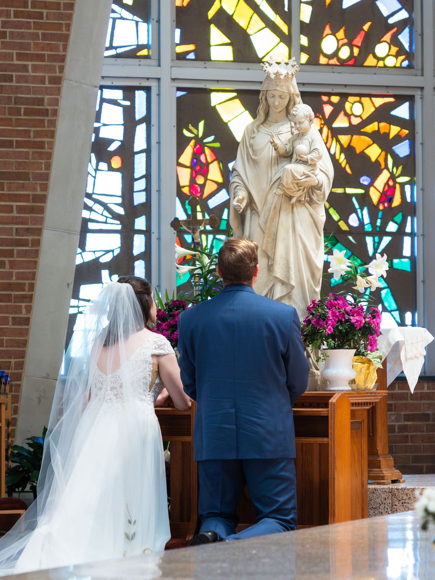A bride and groom kneel before a statue of the Virgin Mary beneath a colorful stained glass window.