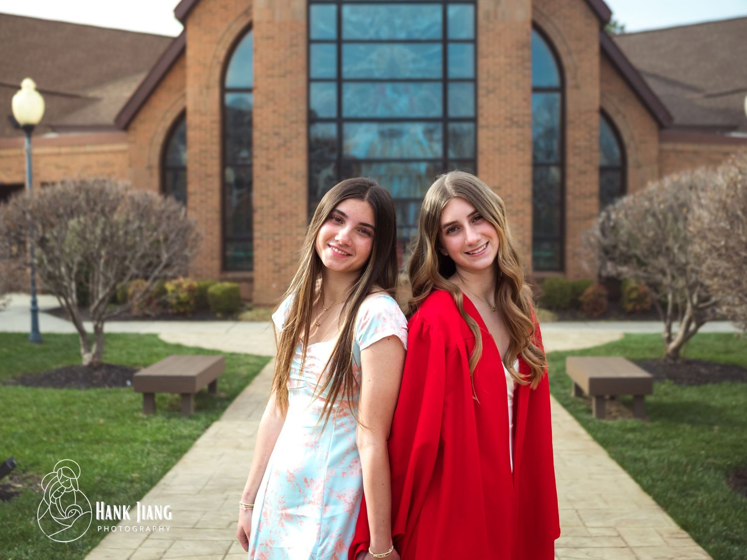 Two teenage girls pose outdoors in front of a brick building, one in a blue dress and one in a red graduation gown.