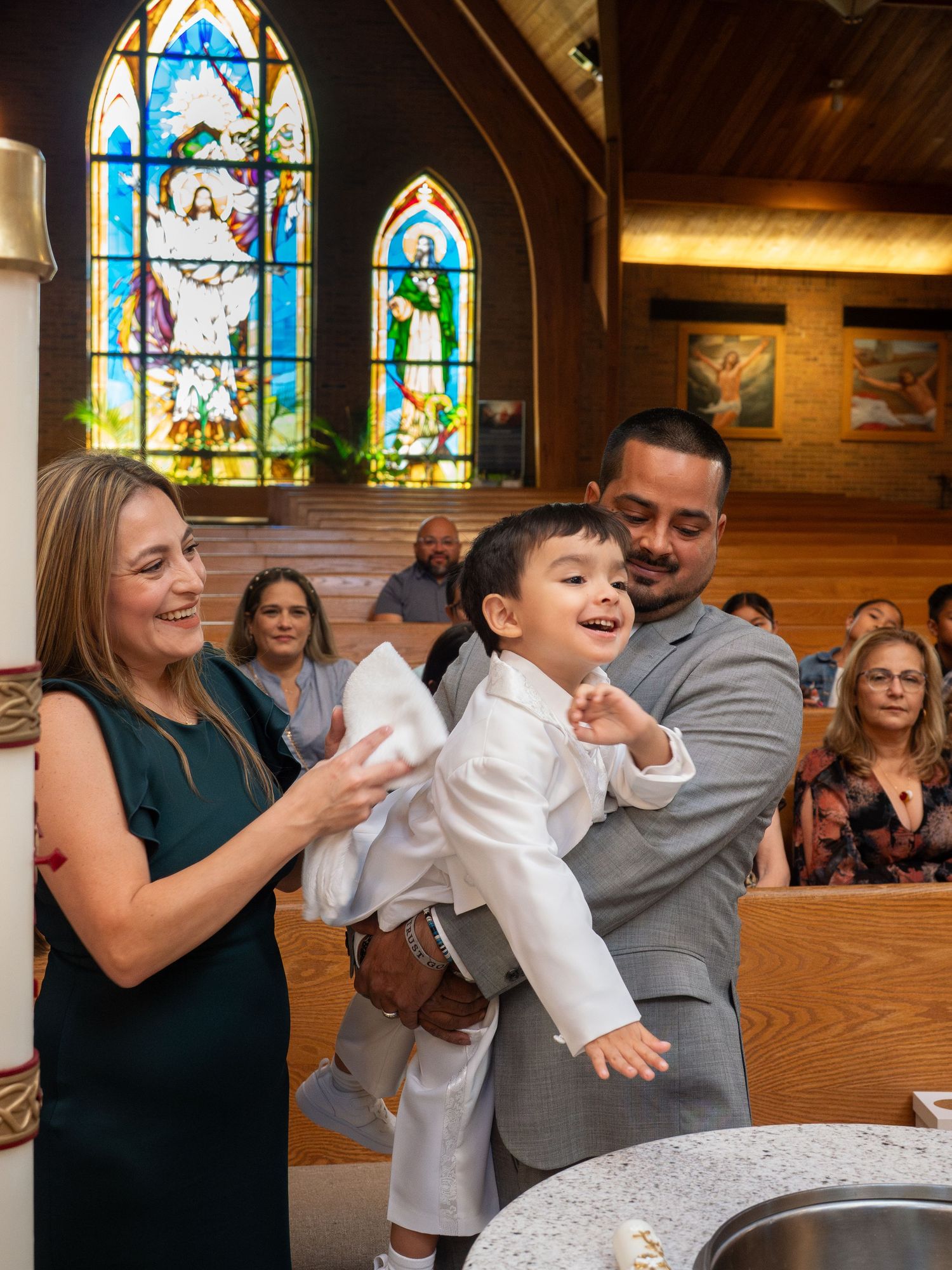 Family celebrating child baptism at Catholic church font with stained glass windows.