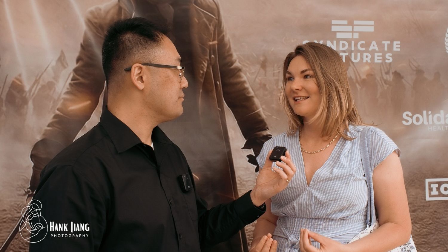 A man in black and a woman in blue dress being interviewed at a Syndicate Pictures event.
