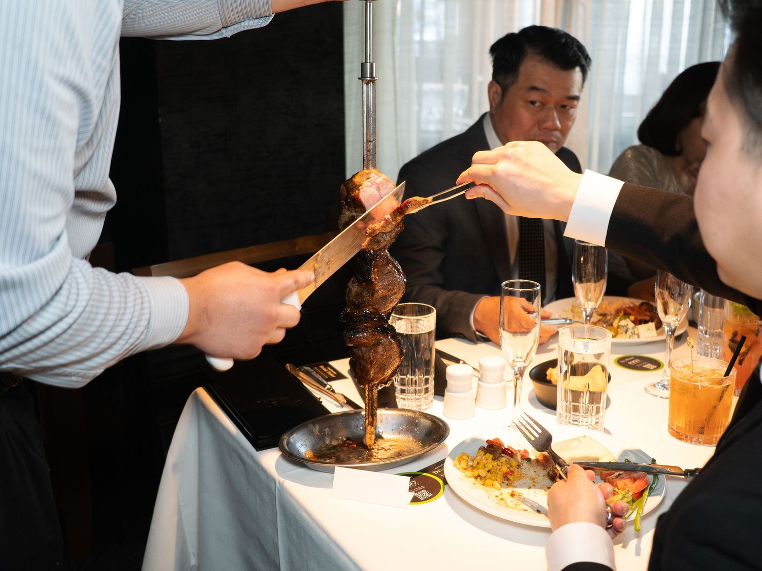 A waiter serves meat with tongs to guests at an elegant restaurant dining table set with glasses and plates.
