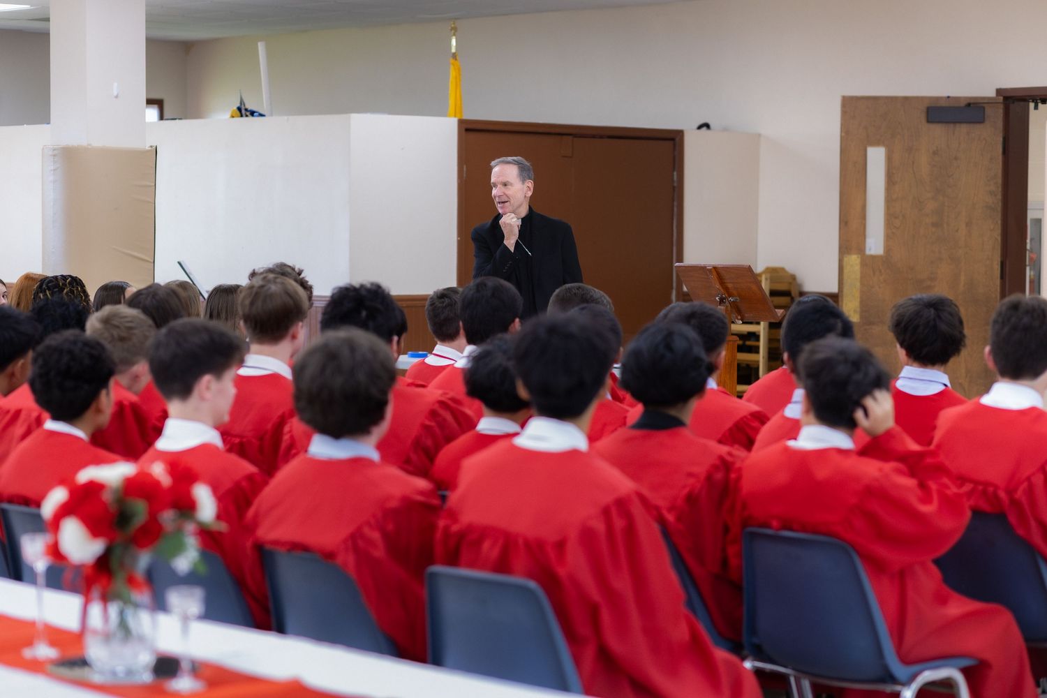 Students in red uniforms attend a formal school assembly led by a speaker at a podium in a bright hall.