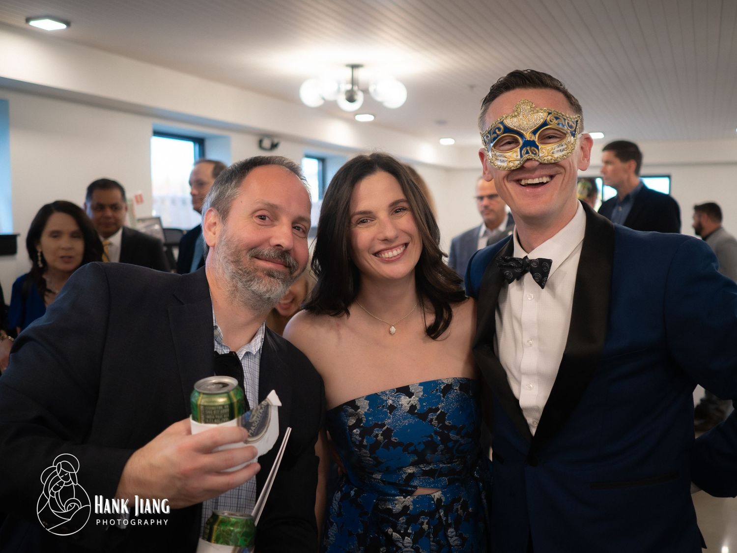 Three guests pose at a masquerade gala, one wearing decorative mask, in formal attire at an elegant event.