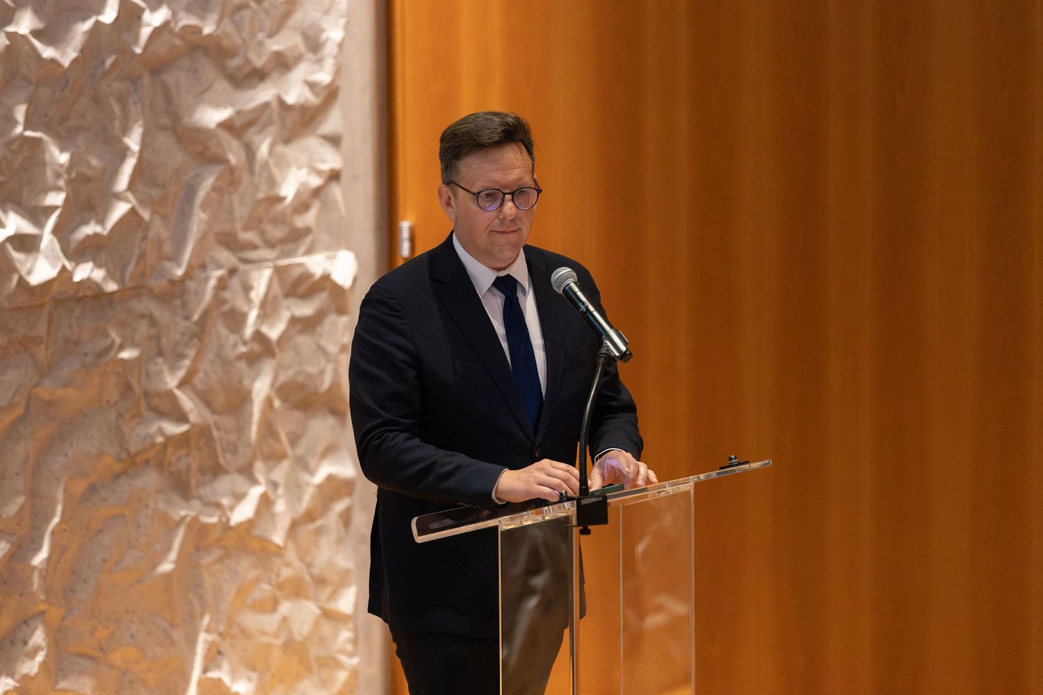 Man in dark suit speaking at a clear podium during a formal event with textured gold wall backdrop.