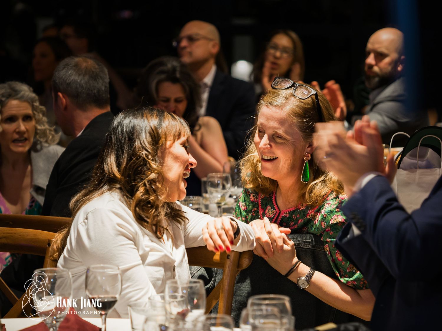 Two women share a joyful moment at a formal dinner event, laughing and holding hands across a table.