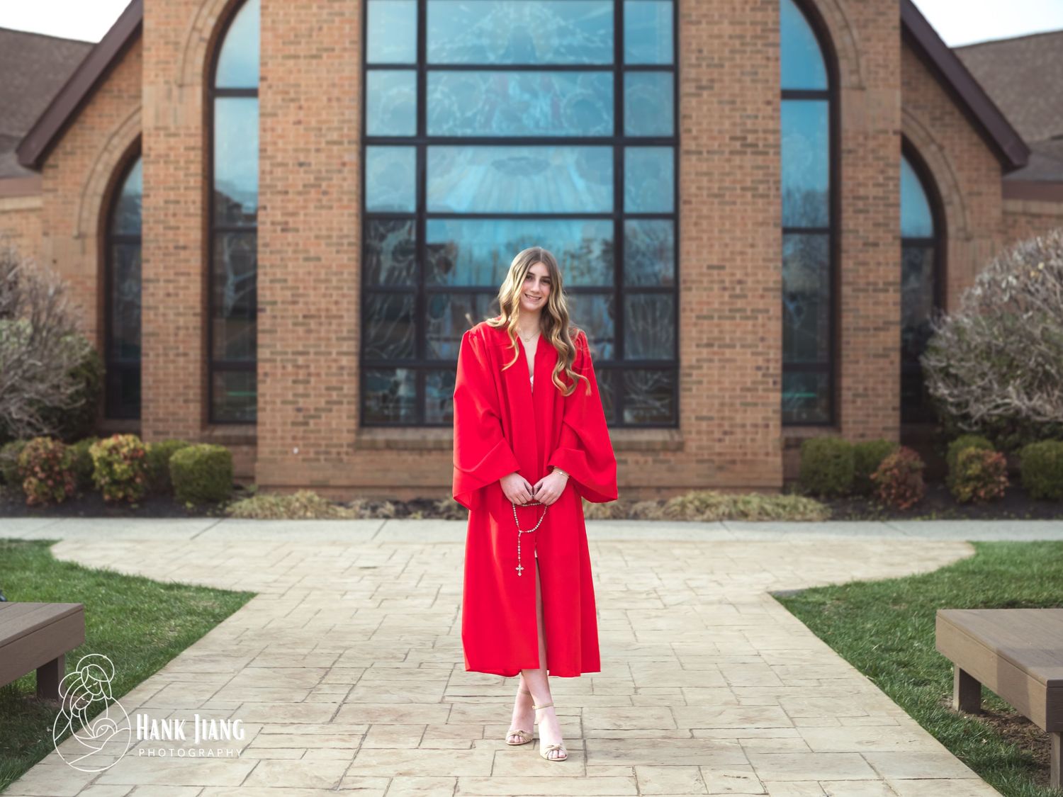 Young woman in red graduation gown smiling outdoors in front of a brick building on a sunny day.