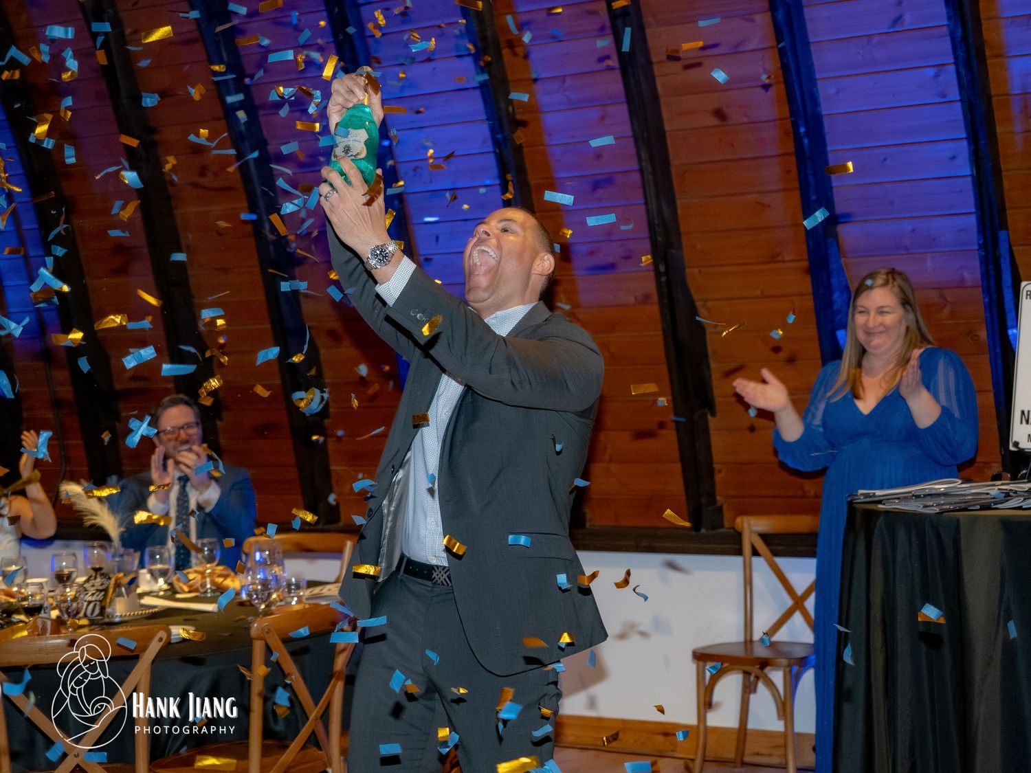 Man celebrates with confetti at an elegant event, raising a bottle while guests cheer in a decorated venue.