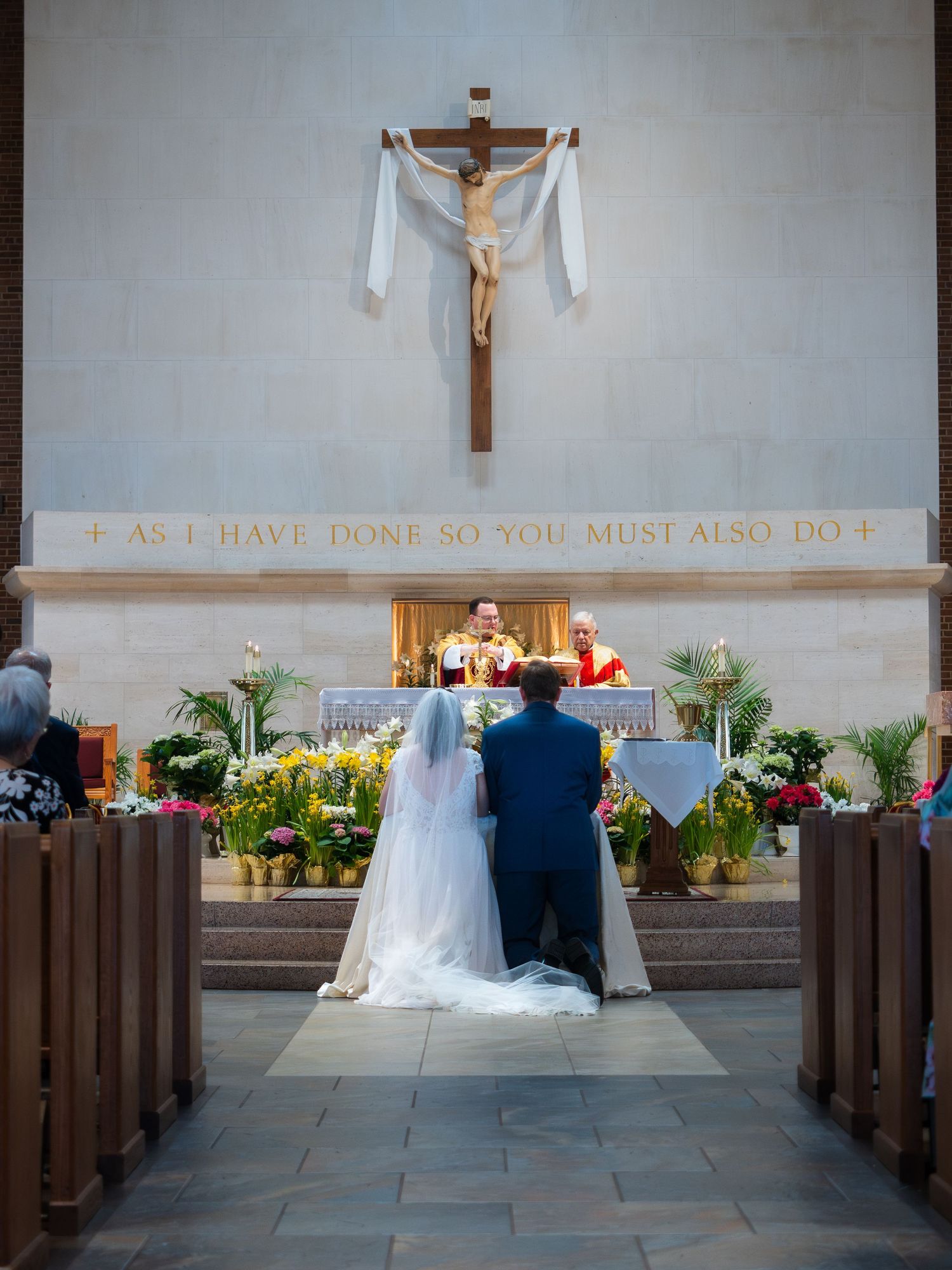 Wedding couple kneeling before priest at altar with crucifix and inscription in Catholic church.