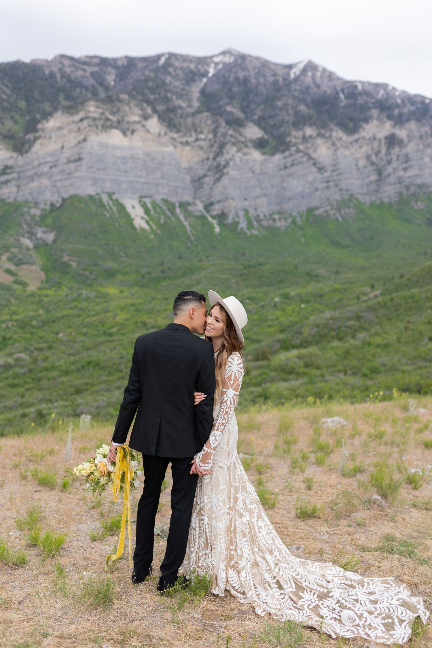 Bride in lace gown and hat with groom standing before a dramatic mountain backdrop in green meadow.