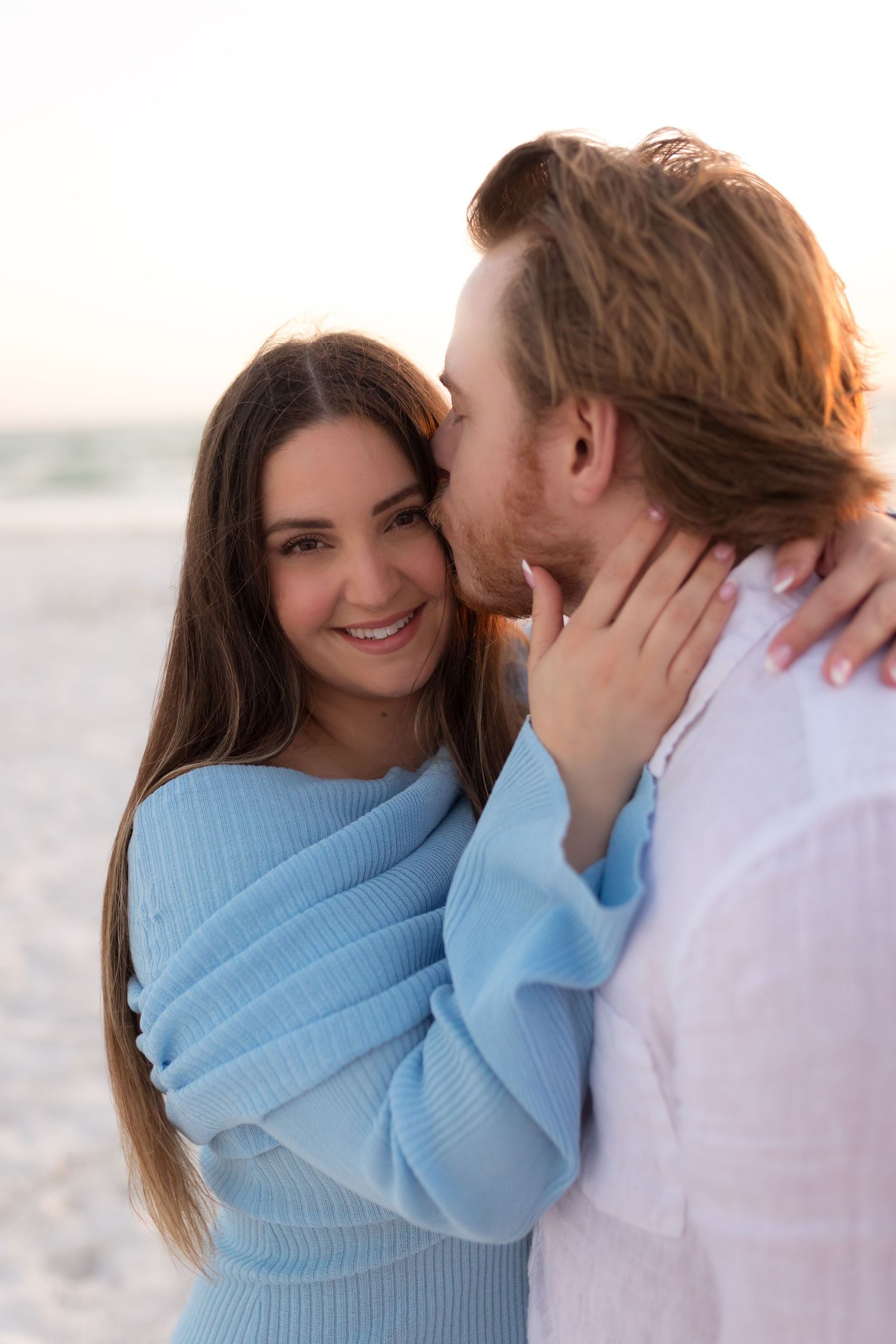 Smiling woman in blue sweater embraces man who kisses her cheek on a sunny beach at sunset.