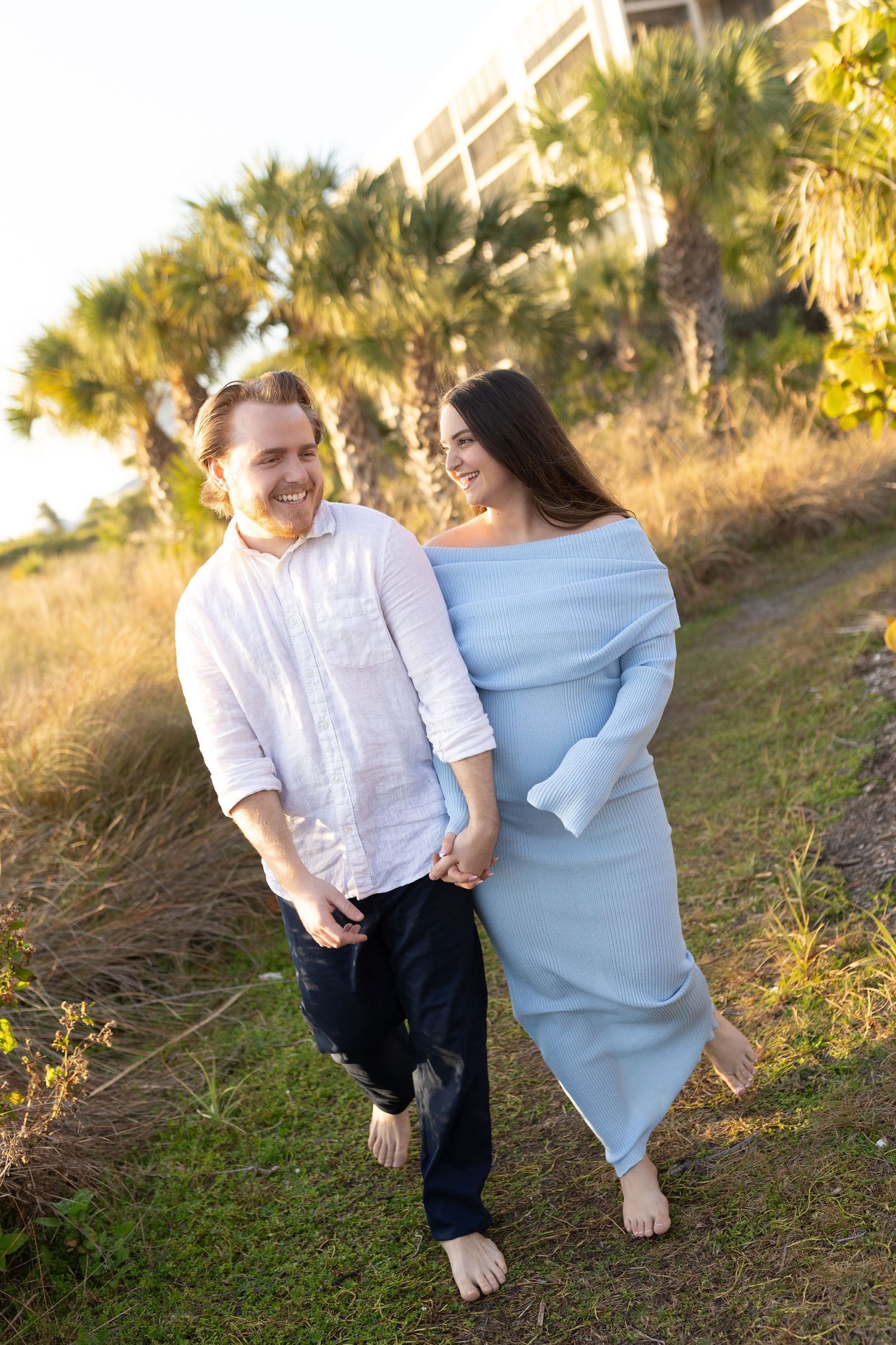 Smiling barefoot couple holding hands and walking together along a coastal path lined with palm trees.