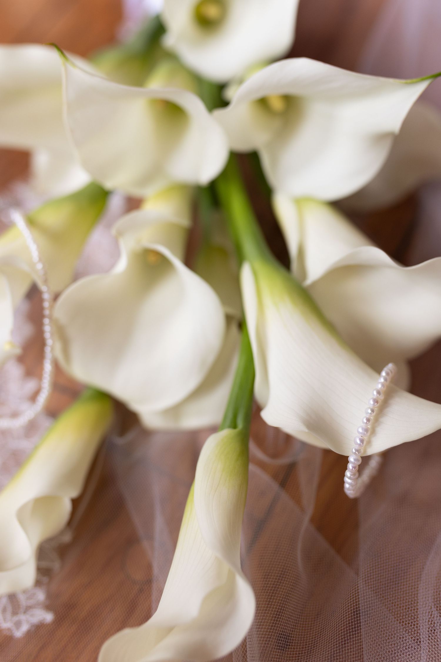 Close-up of elegant white calla lily bridal bouquet with a delicate pearl bracelet on a wooden surface.