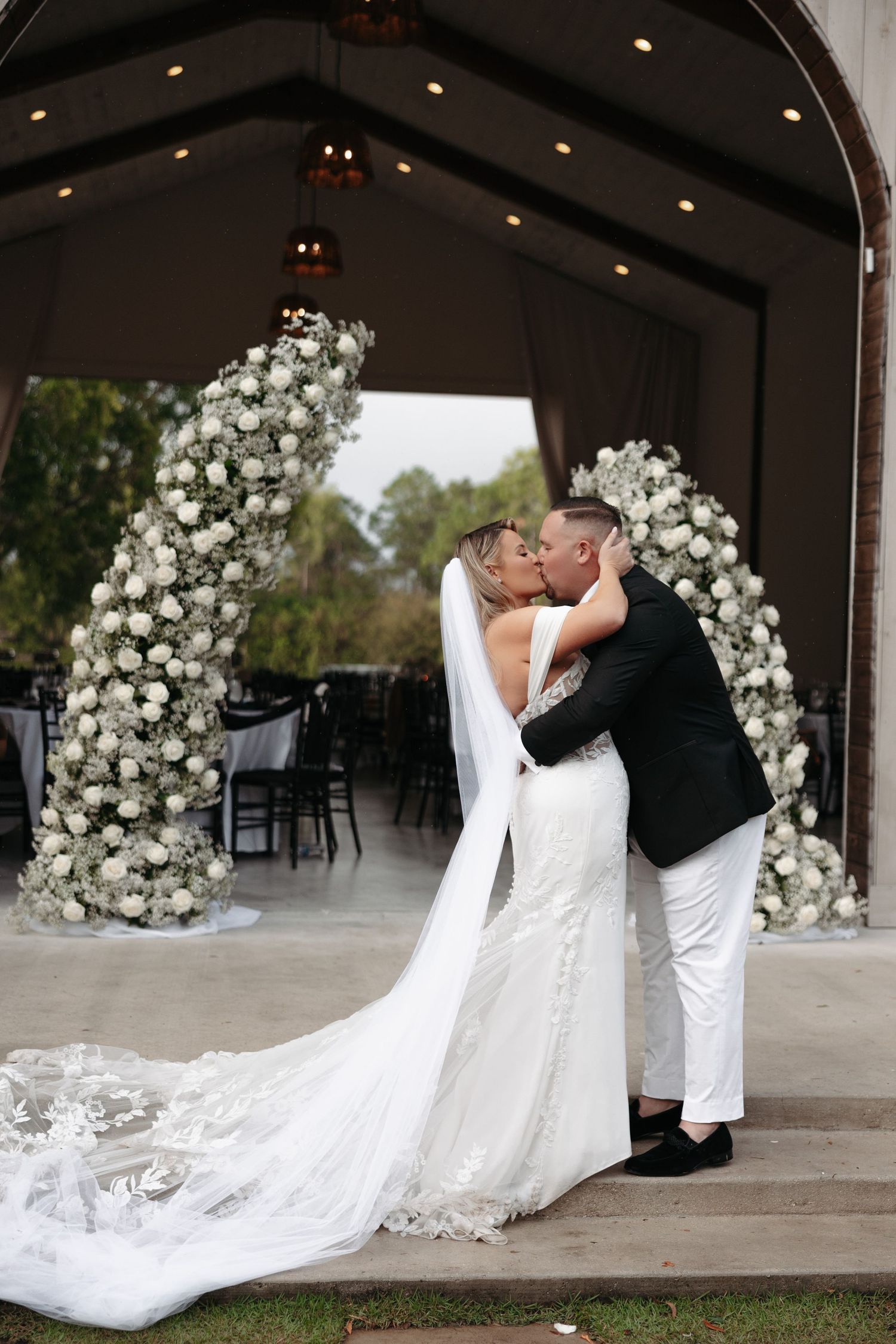 Bride and groom sharing first kiss under floral arch adorned with white roses at outdoor wedding ceremony.