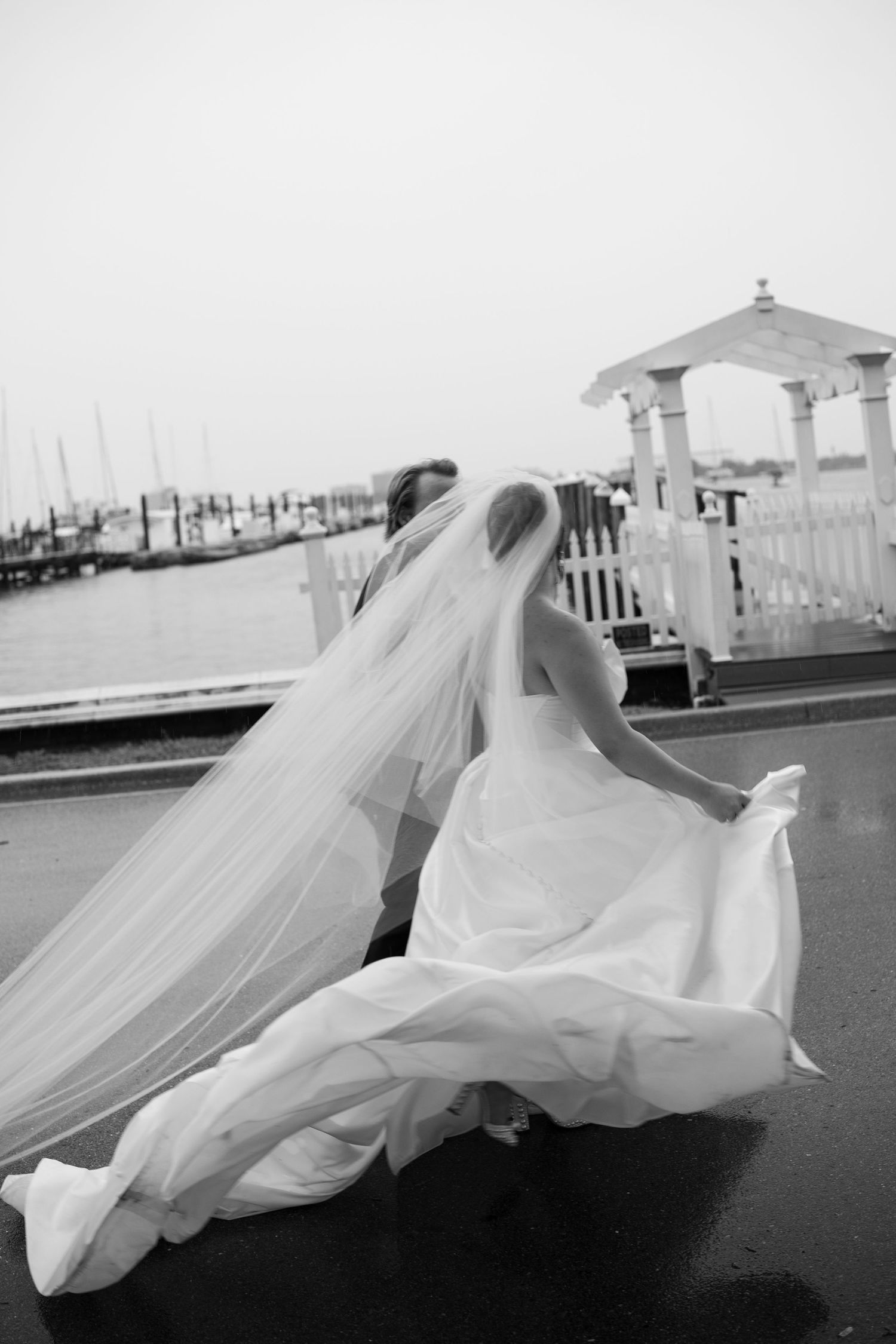 Bride in flowing gown and veil walking near waterfront gazebo in black and white.