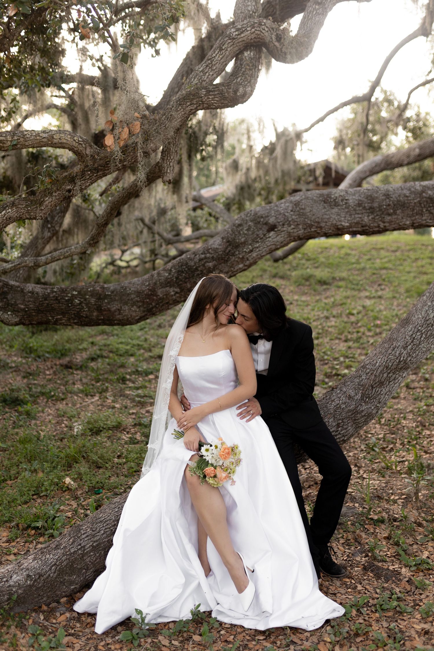 Bride in strapless white gown and groom in black tuxedo sharing a kiss leaning against a large oak tree.