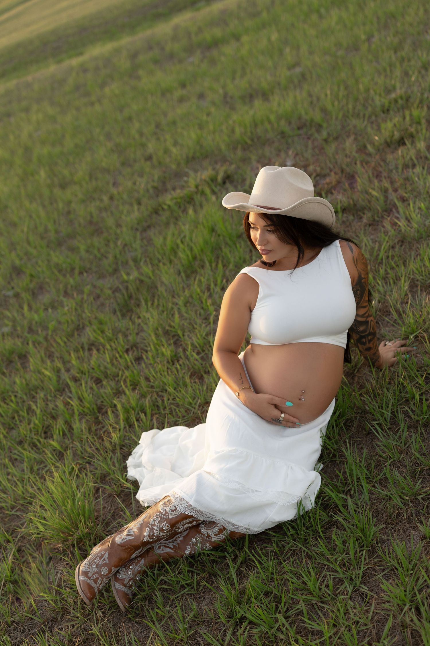 Pregnant woman in white outfit and cowboy hat sits in green field cradling baby bump.