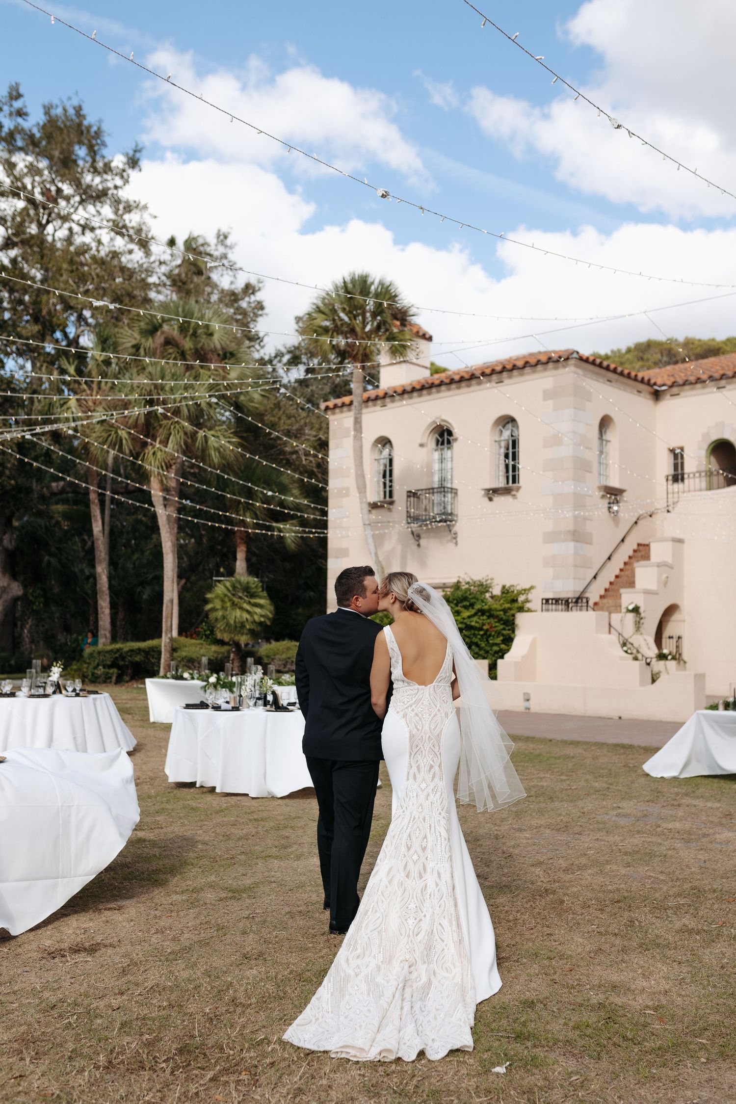 Bride and groom share a kiss at outdoor wedding reception with string lights and Spanish-style venue.
