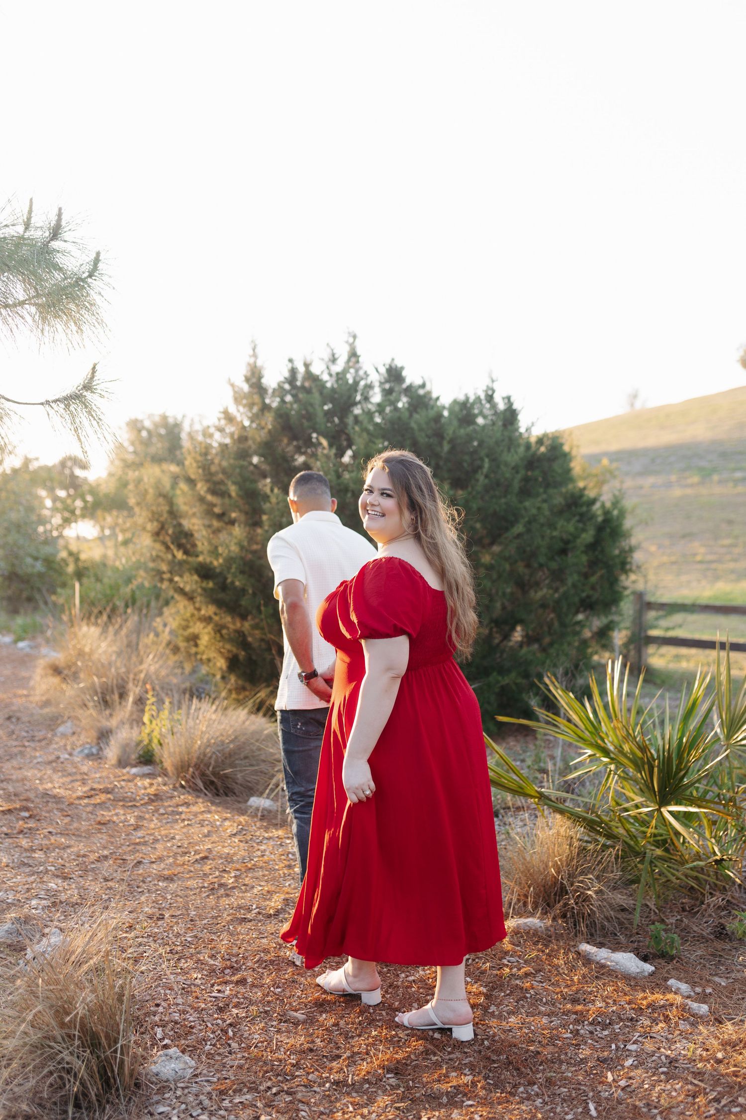 Woman in red dress smiling over shoulder while holding hands with man walking on scenic trail.