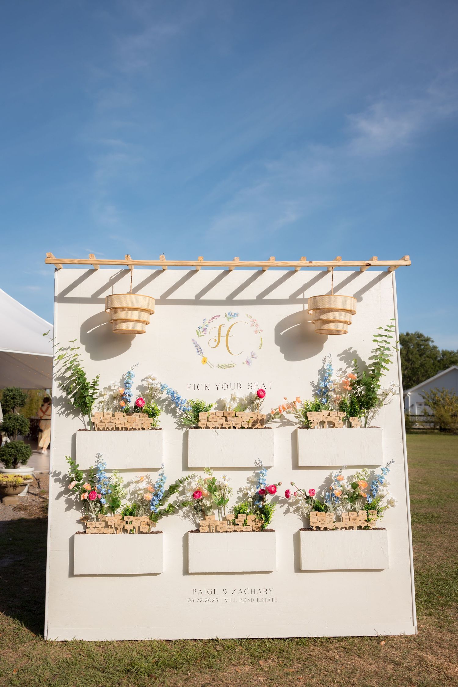 Outdoor wedding seating chart display with floral arrangements on a white pergola wall.