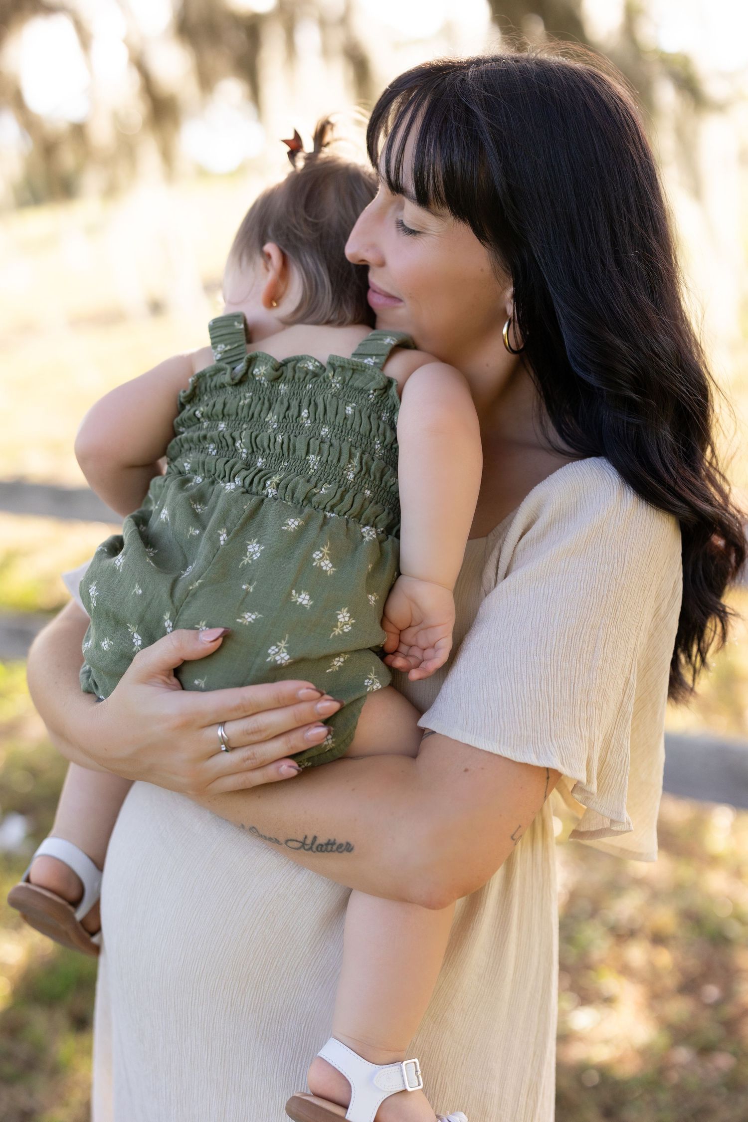 Dark-haired mother tenderly holds sleeping baby in green floral outfit outdoors in soft light.