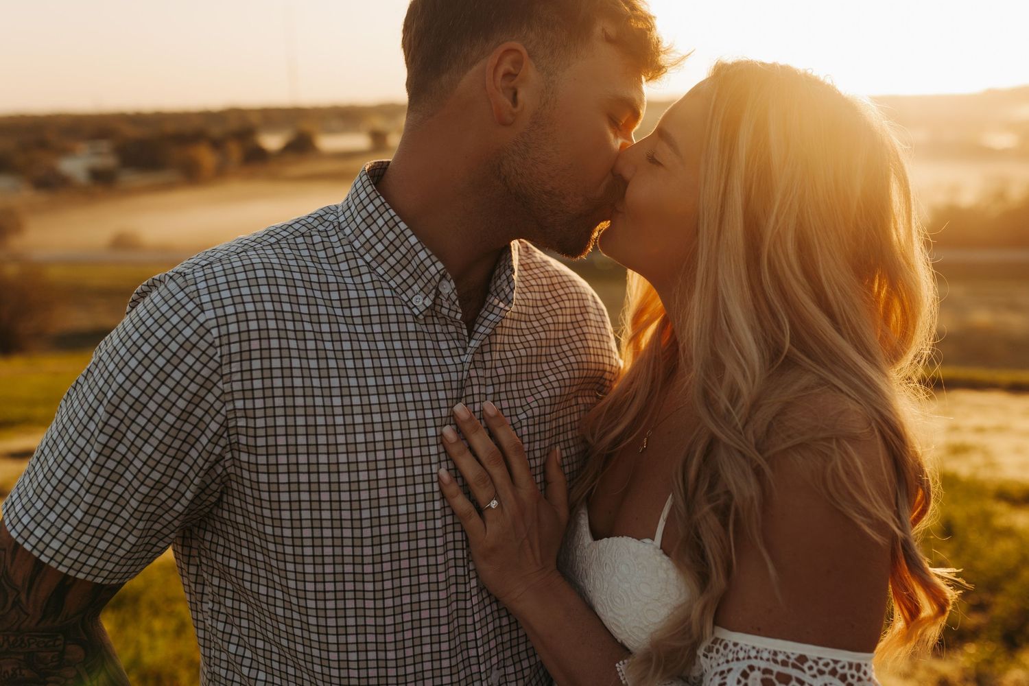 Couple sharing a tender kiss outdoors in warm golden sunset light in a scenic countryside setting.