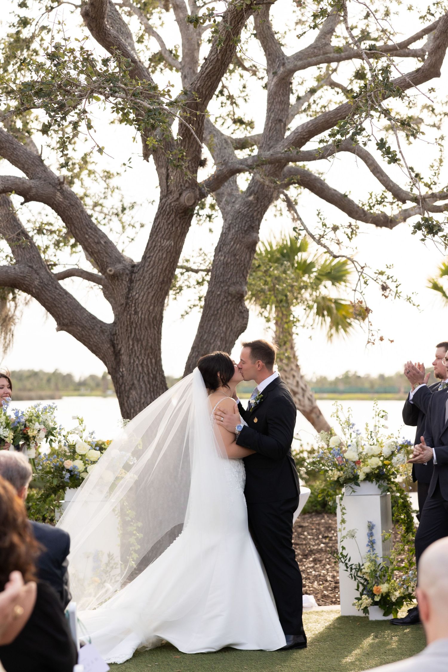 Bride and groom share first kiss under a grand oak tree during outdoor wedding ceremony.