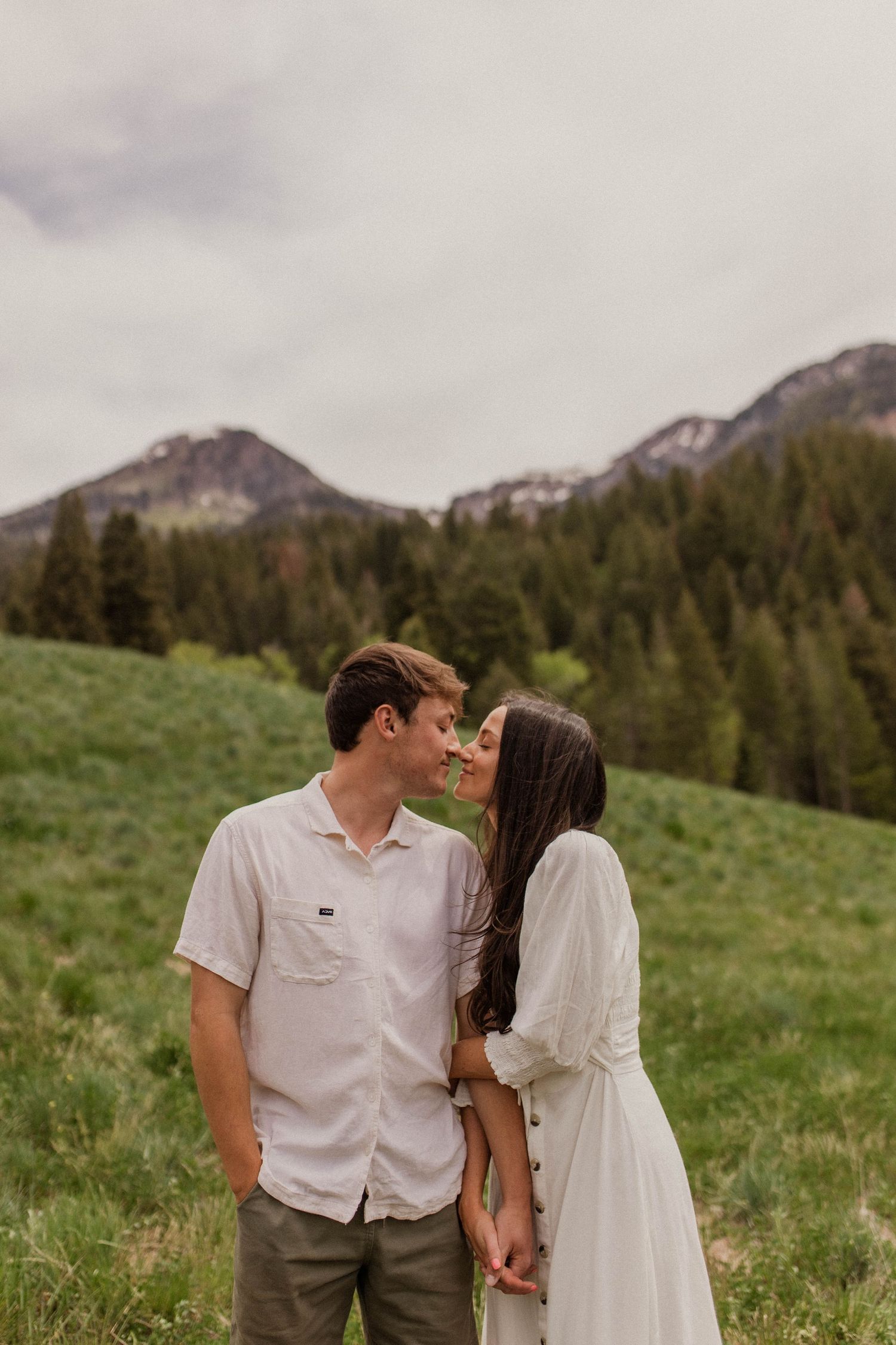 Couple sharing a tender almost-kiss in a green mountain meadow with forested peaks in the background.