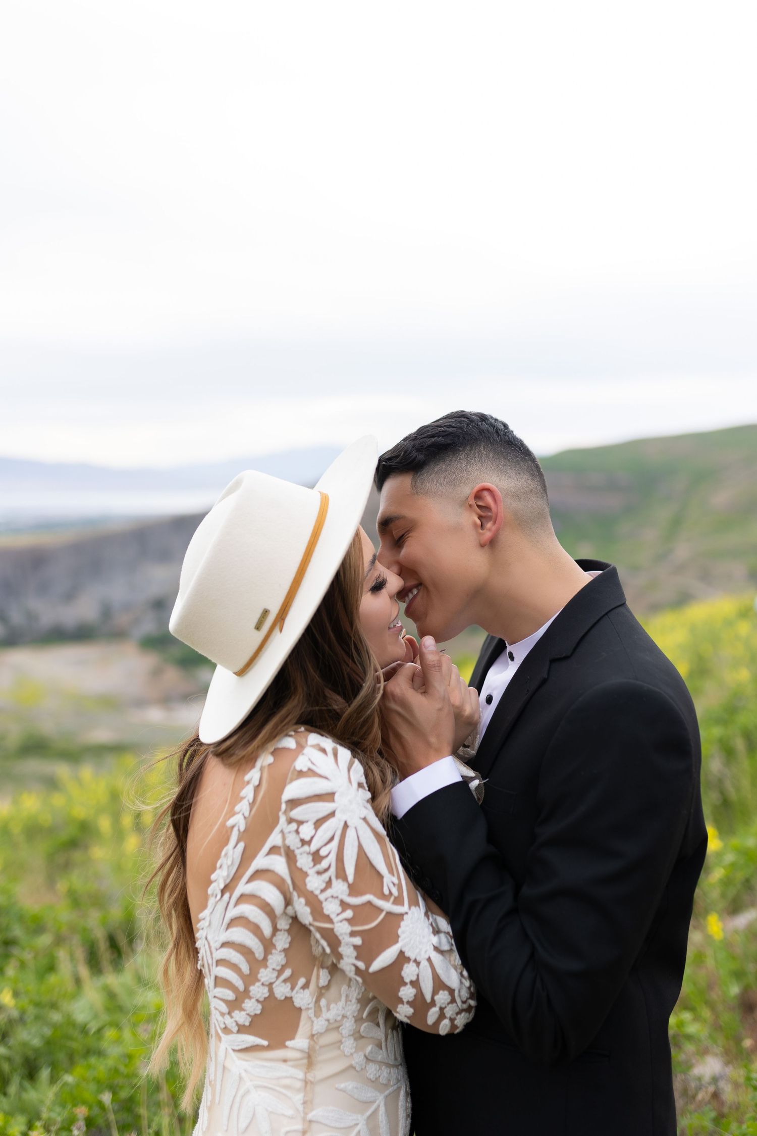 Couple sharing an intimate moment, bride in lace gown and wide-brim hat, surrounded by mountain scenery.