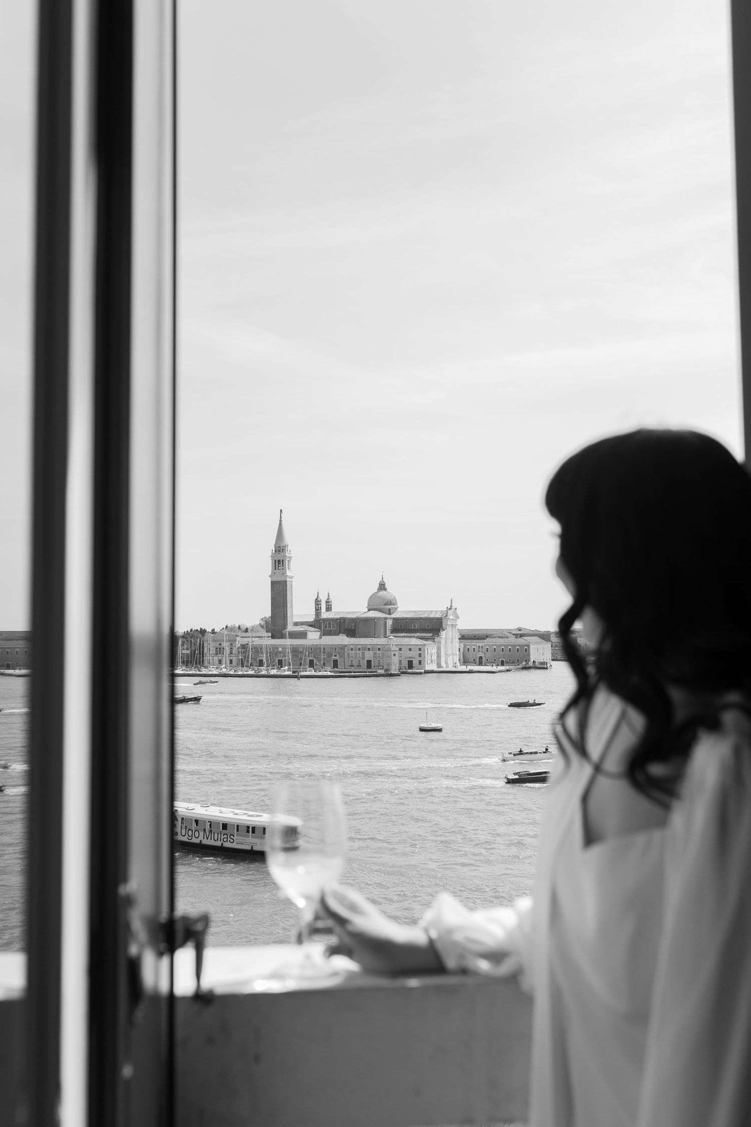Black and white photo of a bride gazing at the Venice lagoon and San Giorgio Maggiore from a window.