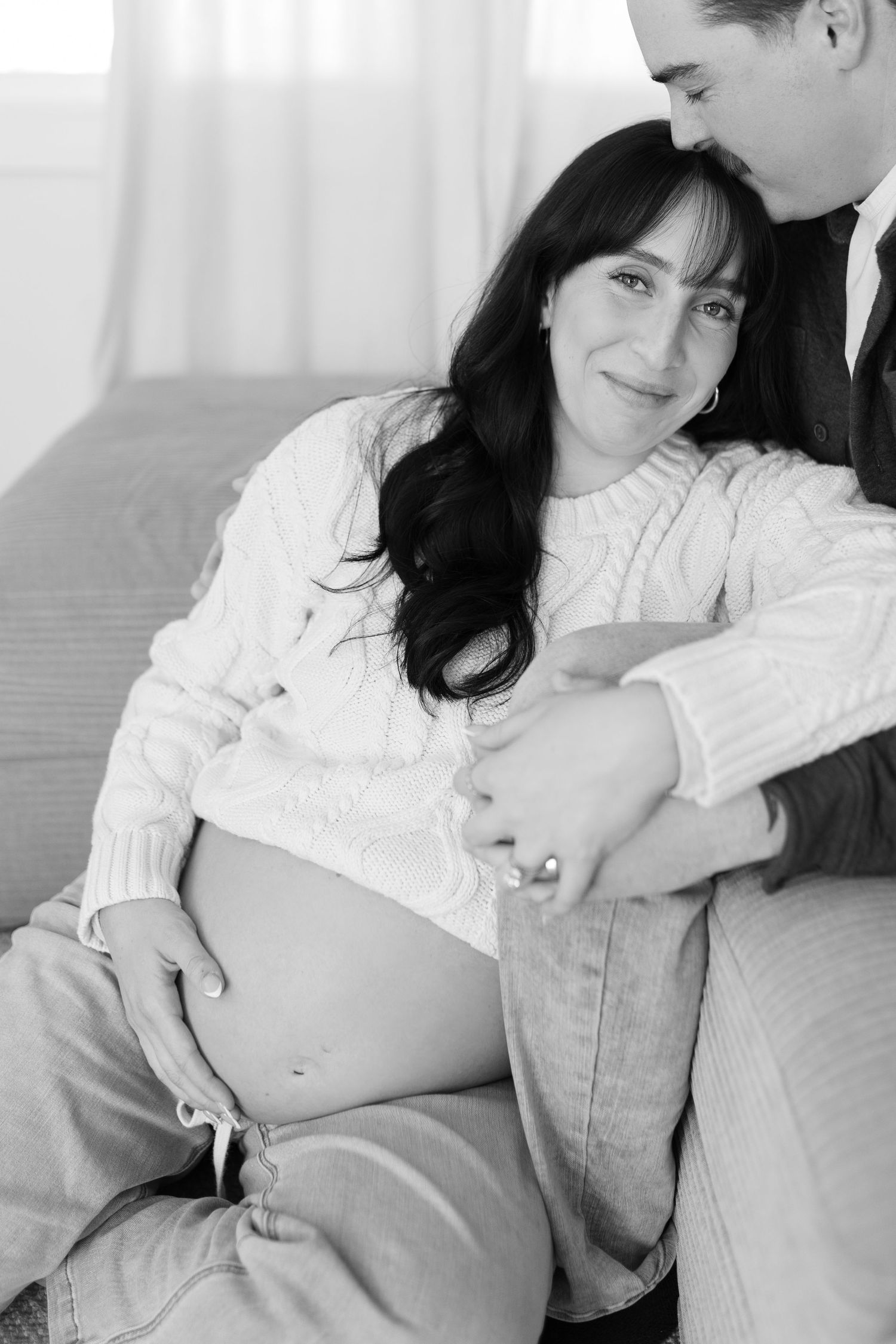 Black and white maternity portrait of pregnant woman sitting between partners legs on couch.
