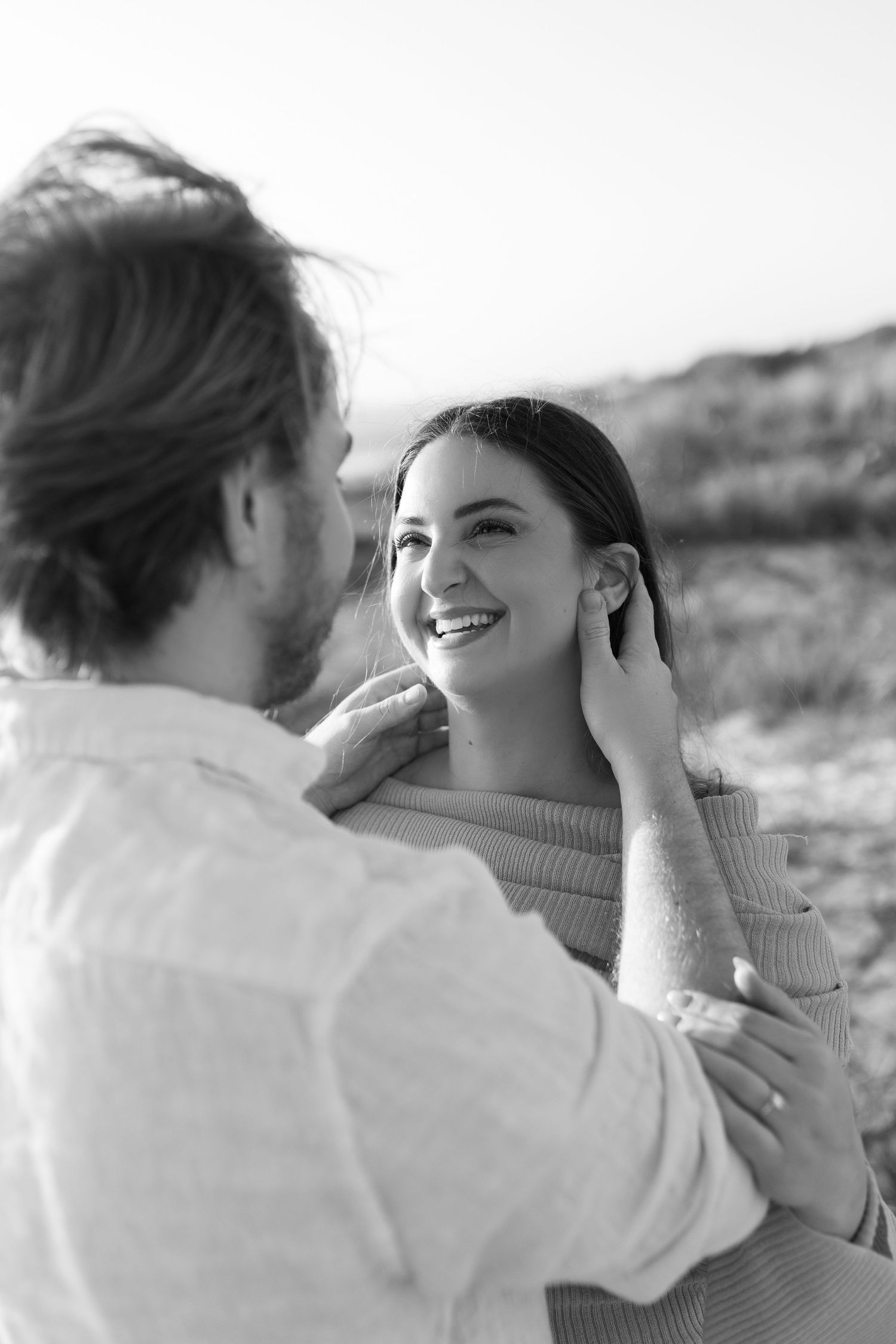 Black and white photo of laughing woman as man gently touches her face among sandy beach dunes.