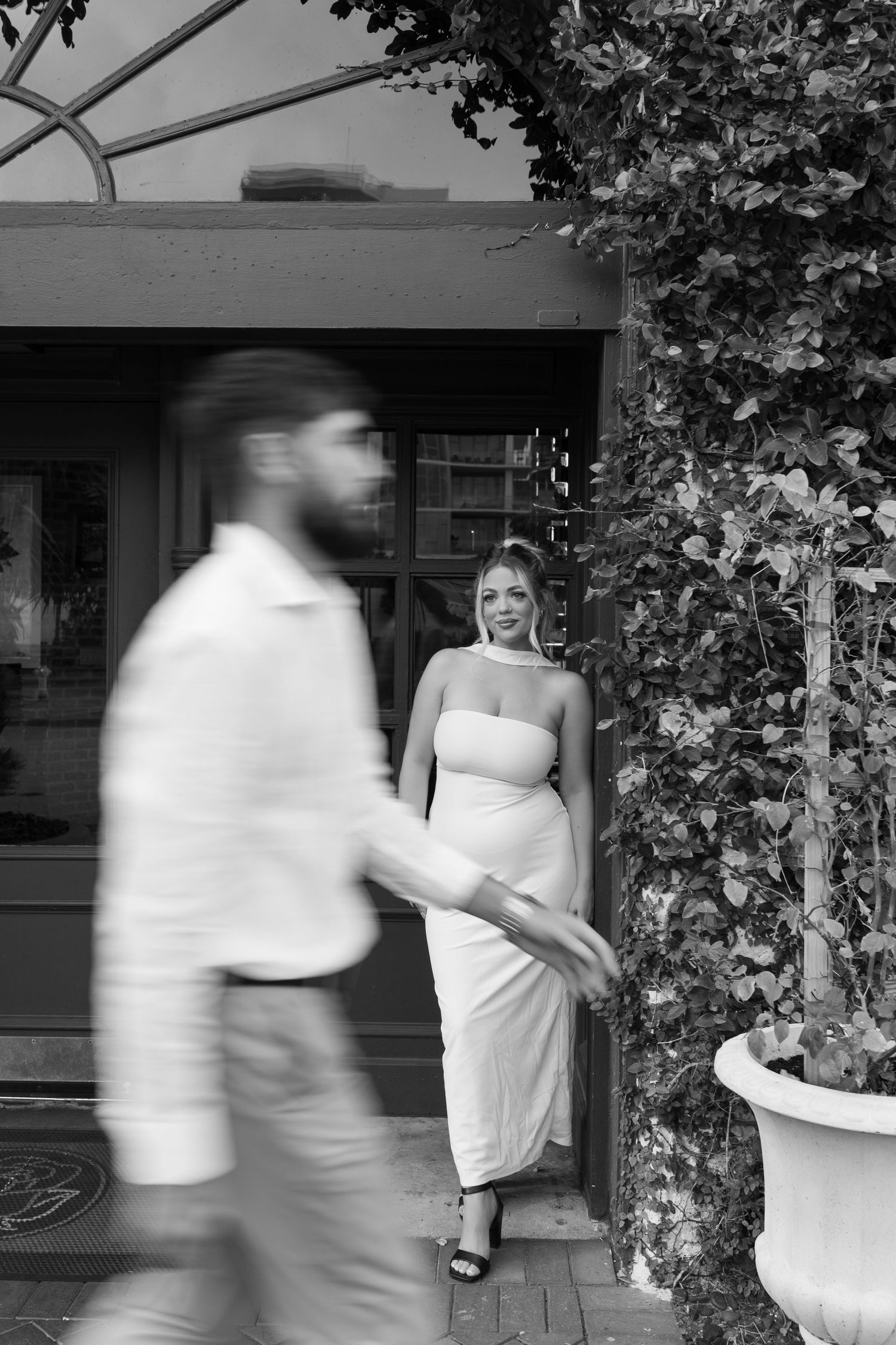 Black and white photo of a woman in white dress posing by ivy-covered doorway while man walks past in foreground.