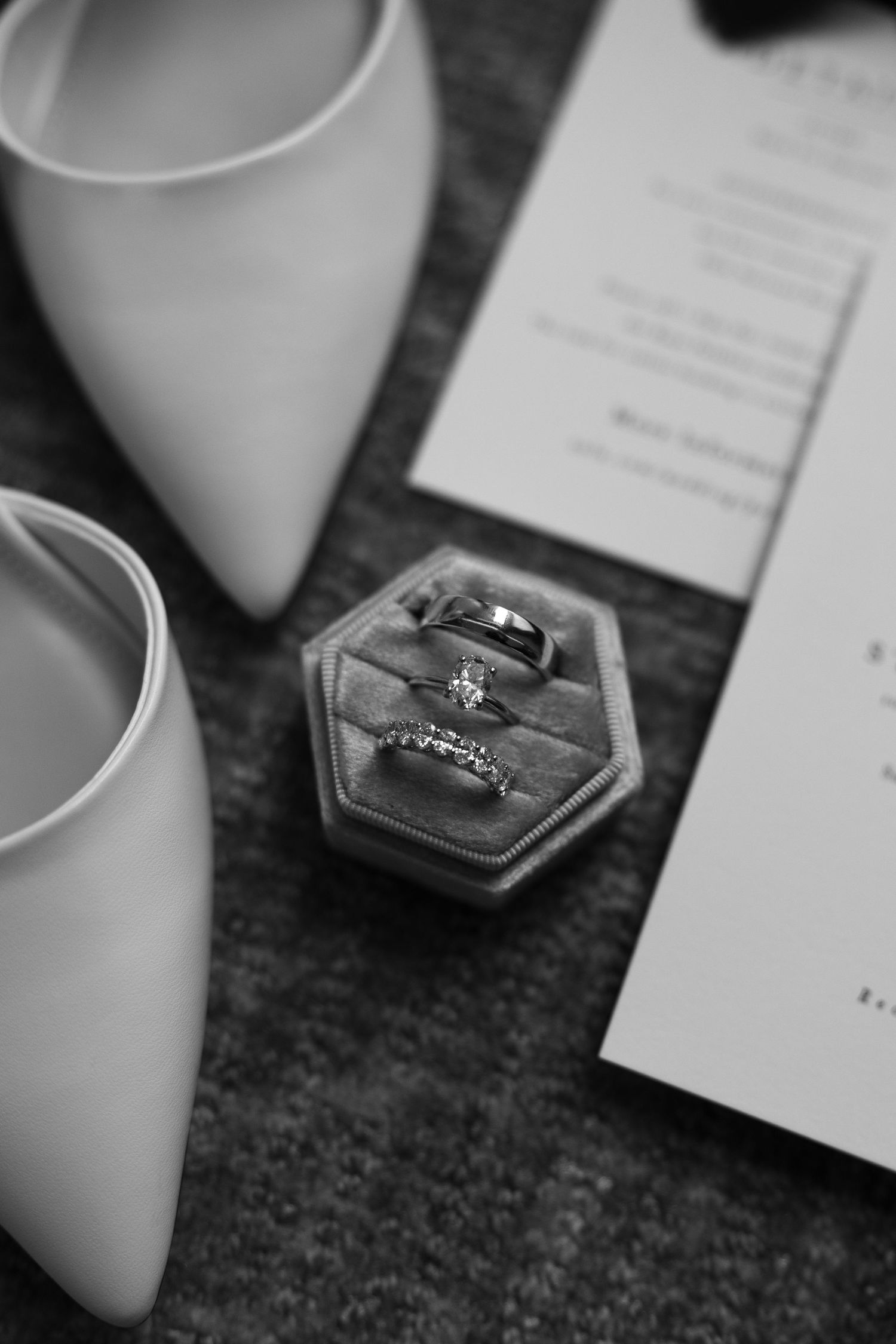 Black and white photo of wedding rings in a velvet hexagon box beside white bridal heels and invitation.